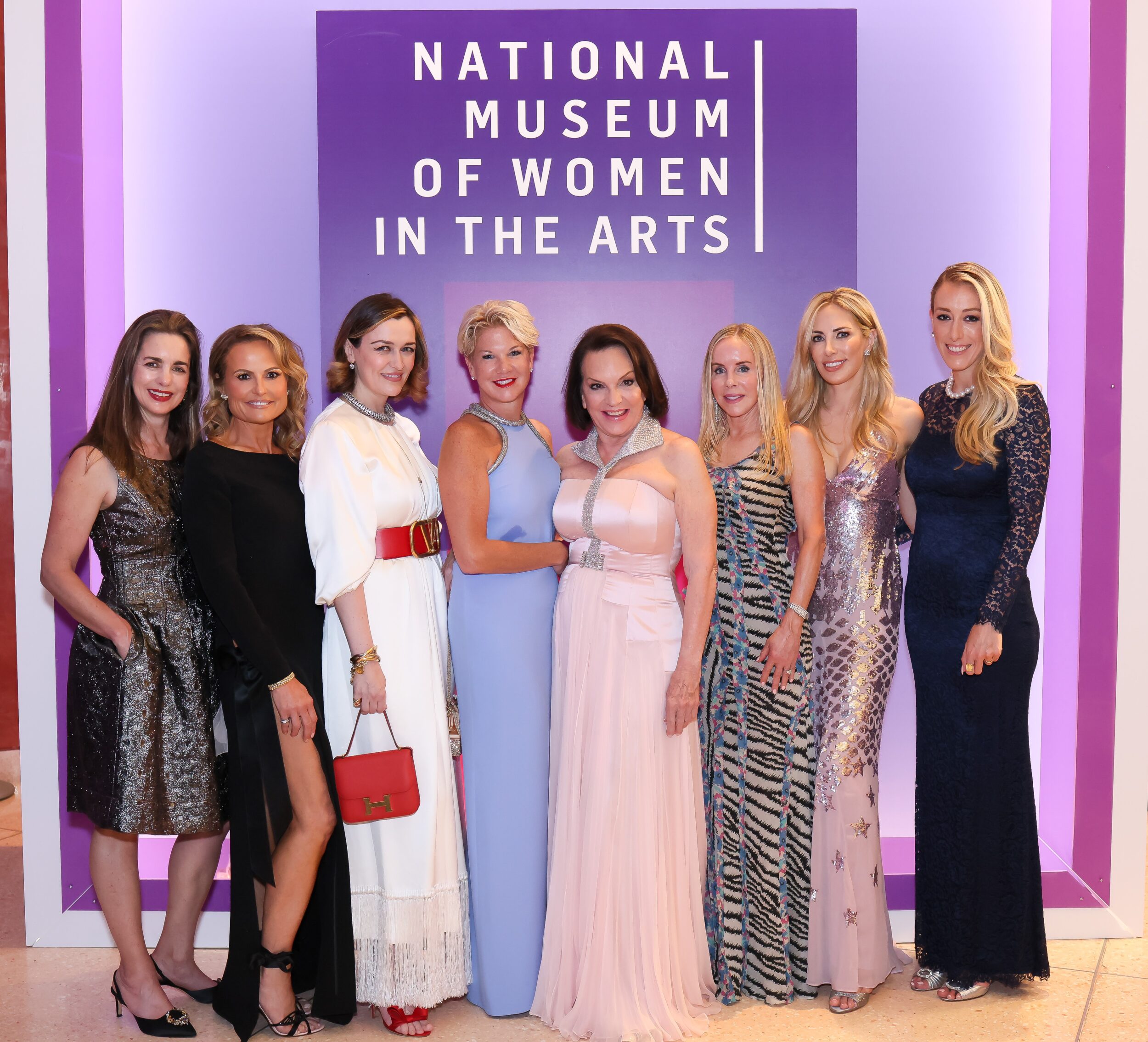 A group of light-skinned women pose for a photo at a black-tie event. Behind the women is a colorful backdrop with the National Museum of Women in the Arts logo on it.
