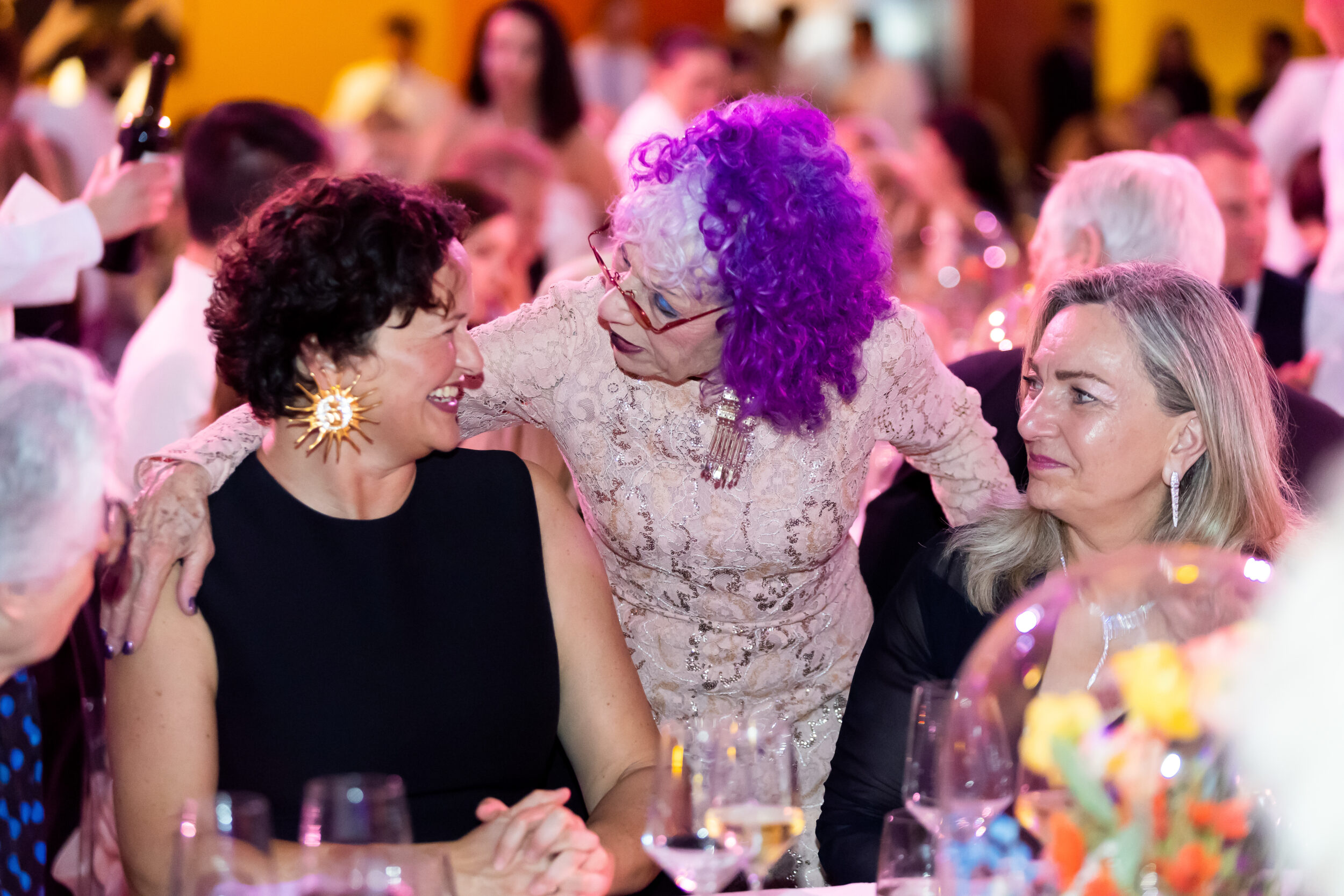 A light-skinned woman with purple hair leans down to chat with two light-skinned women seated at a black-tie event.