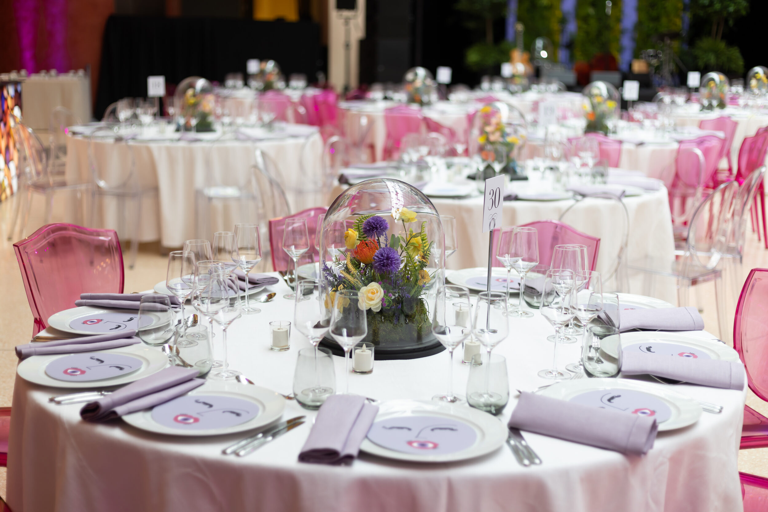 A table setting for a black-tie event with pink acrylic chairs and a small colorful flower arrangement under a glass cloche.