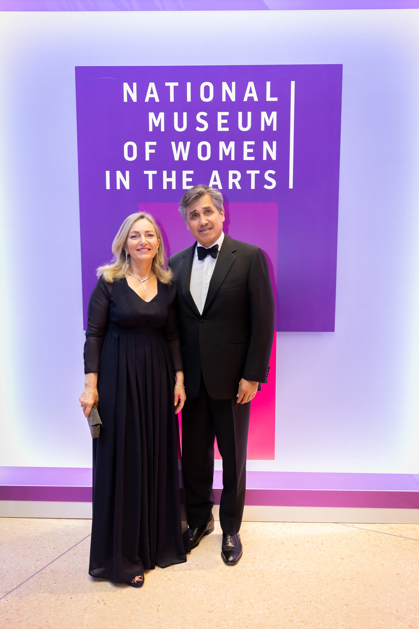 A light-skinned man and woman pose for a photo at a black-tie event. Behind the couple is a colorful backdrop with the National Museum of Women in the Arts logo on it.