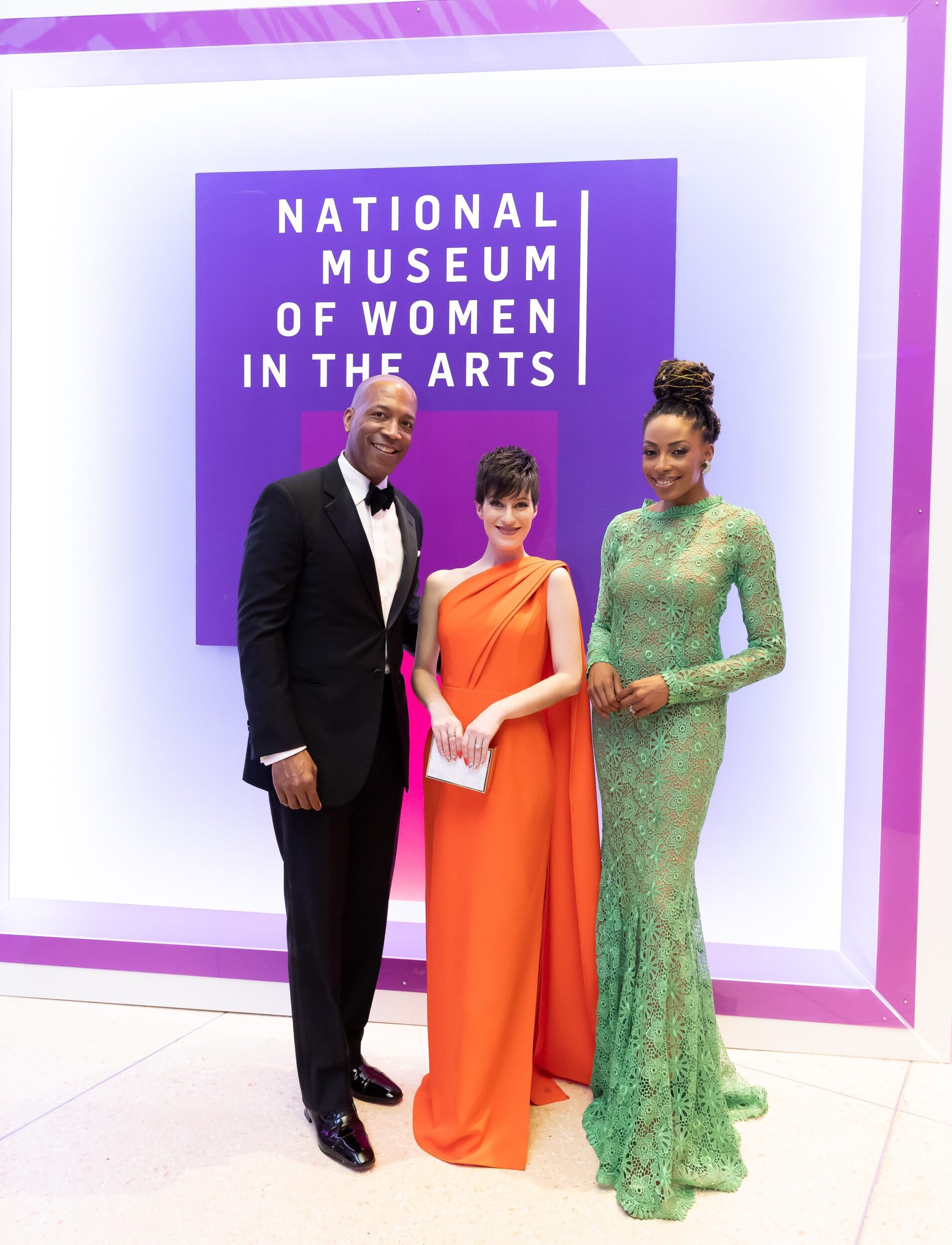 A medium-skinned man and woman pose for a photo with a light-skinned woman at a black-tie event. Behind the group is a colorful backdrop with the National Museum of Women in the Arts logo on it.