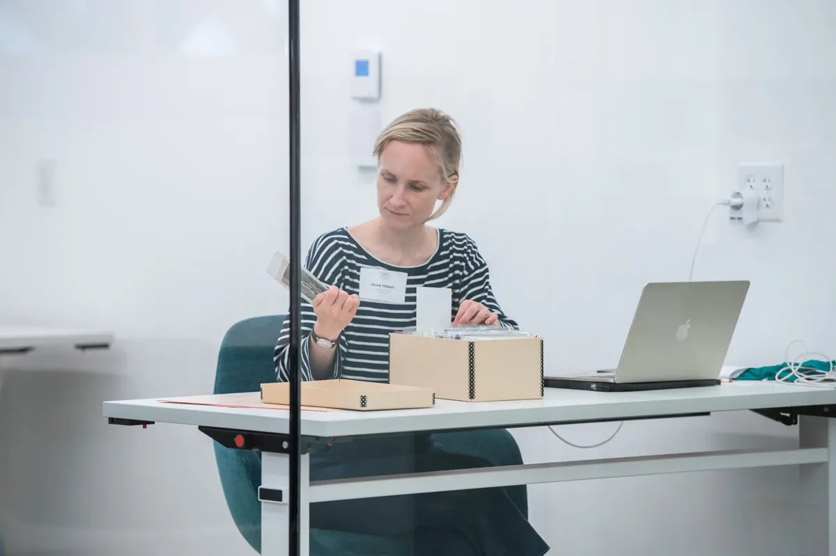 A woman with a light skin tone and blonde hair is sitting behind a glass wall by a desk. With one hand, she is holding a card and taking a closer look, with the other hand she is holding onto a box of cards.