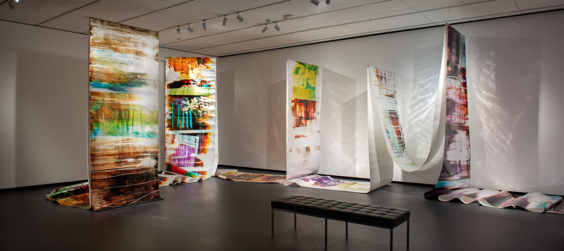View of a gallery space. Several large tapestries are hanging from the ceiling. Colorful broad paint strokes are painted onto the white canvases. The canvases are partly covering the floor. A bench is standing in the middle of the gallery.