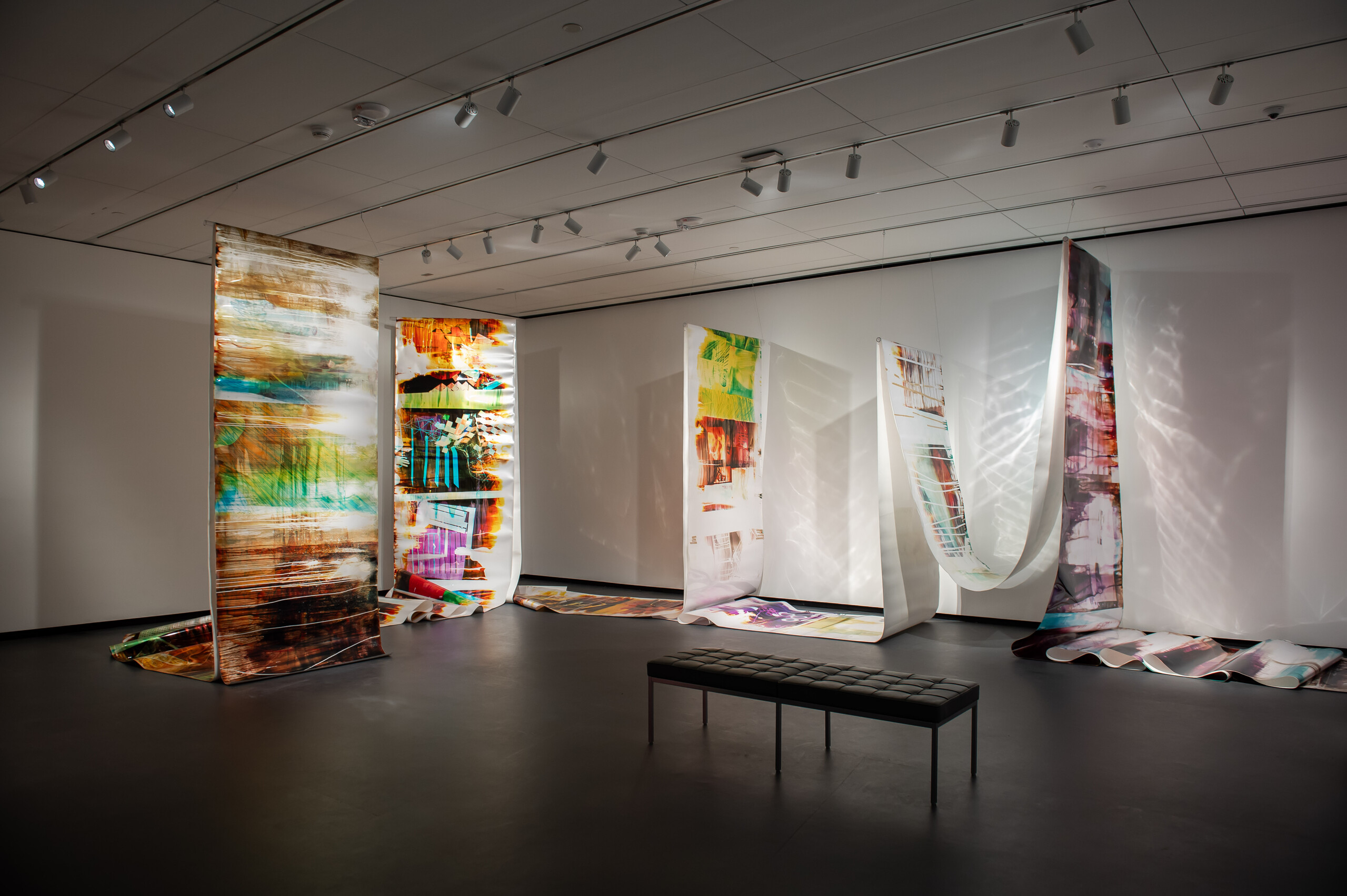 View of a gallery space. Several large tapestries are hanging from the ceiling. Colorful broad paint strokes are painted onto the white canvases. The canvases are partly covering the floor. A bench is standing in the middle of the gallery.