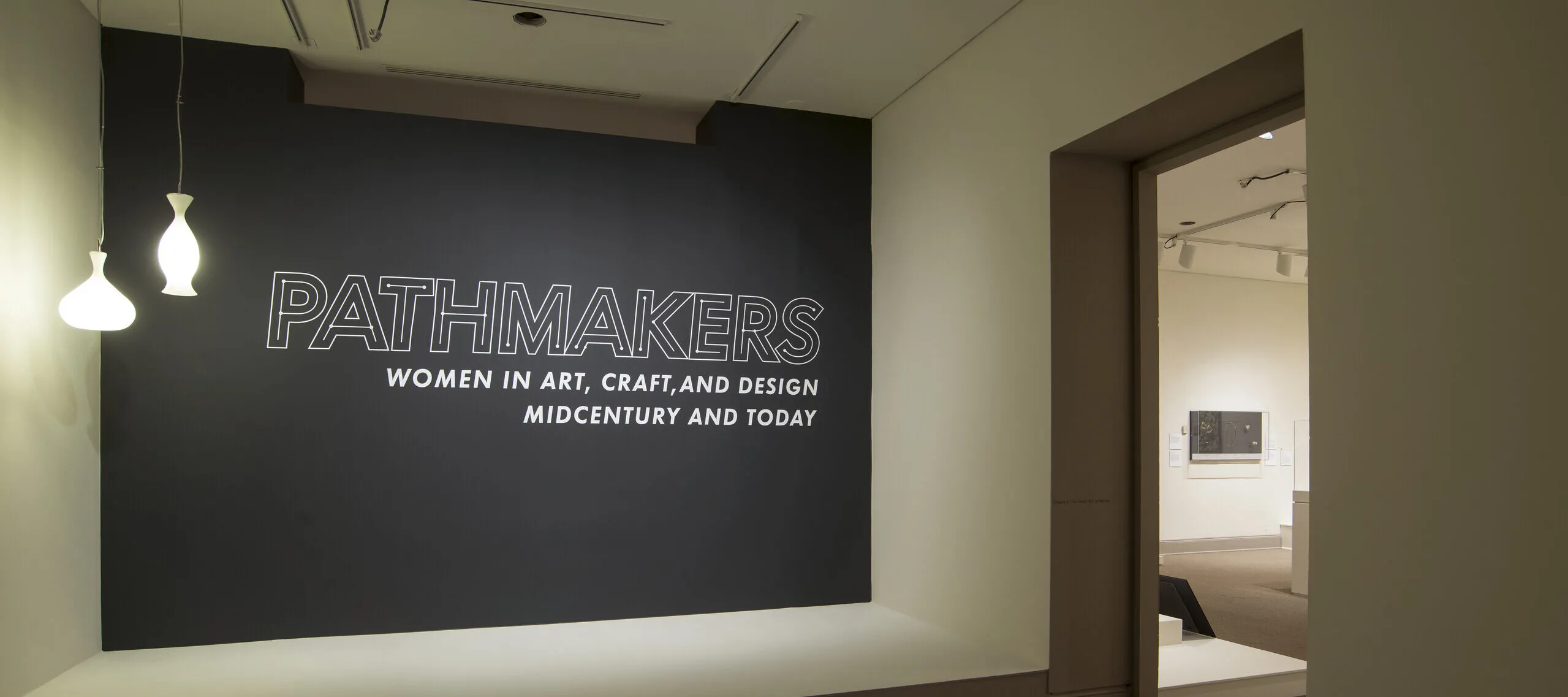 View of a gallery space. On a black wall, it says " Pathmakers: Women in Art, Craft, and Design, Midcentury and Today" in big, white letters, Two pendant lights are hanging from the ceiling to the left.