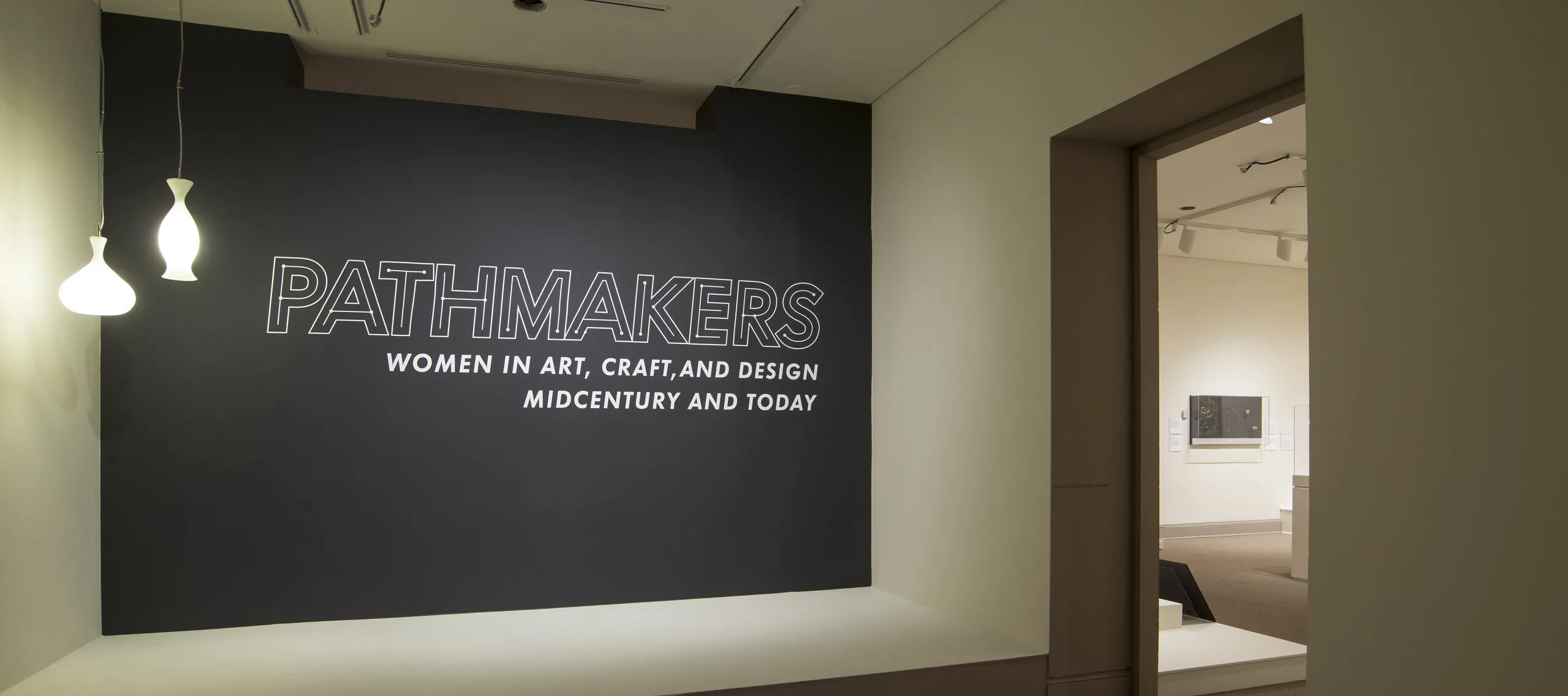 View of a gallery space. On a black wall, it says " Pathmakers: Women in Art, Craft, and Design, Midcentury and Today" in big, white letters, Two pendant lights are hanging from the ceiling to the left.