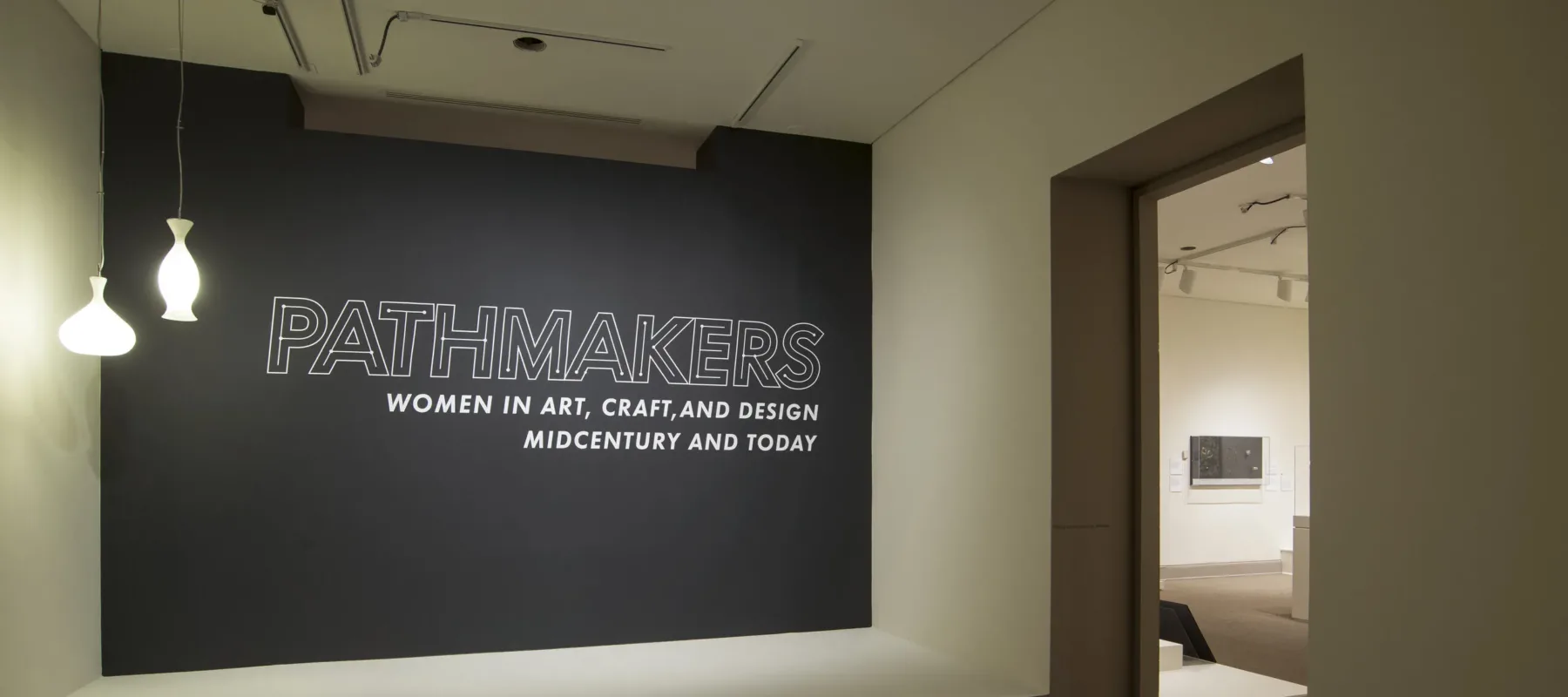 View of a gallery space. On a black wall, it says " Pathmakers: Women in Art, Craft, and Design, Midcentury and Today" in big, white letters, Two pendant lights are hanging from the ceiling to the left.