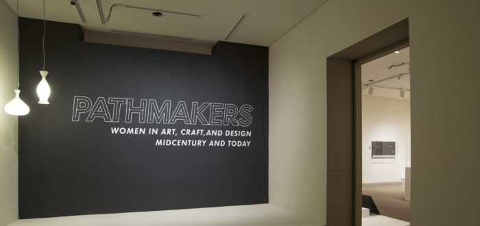 View of a gallery space. On a black wall, it says " Pathmakers: Women in Art, Craft, and Design, Midcentury and Today" in big, white letters, Two pendant lights are hanging from the ceiling to the left.