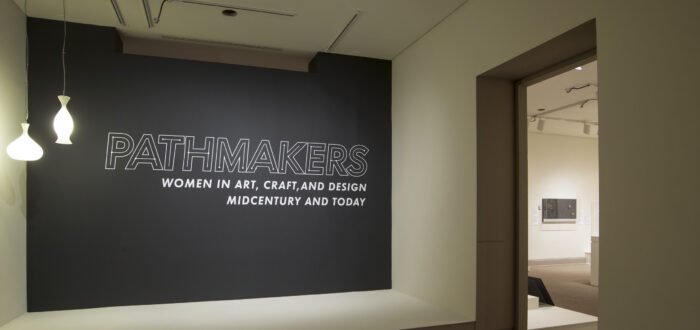 View of a gallery space. On a black wall, it says " Pathmakers: Women in Art, Craft, and Design, Midcentury and Today" in big, white letters, Two pendant lights are hanging from the ceiling to the left.