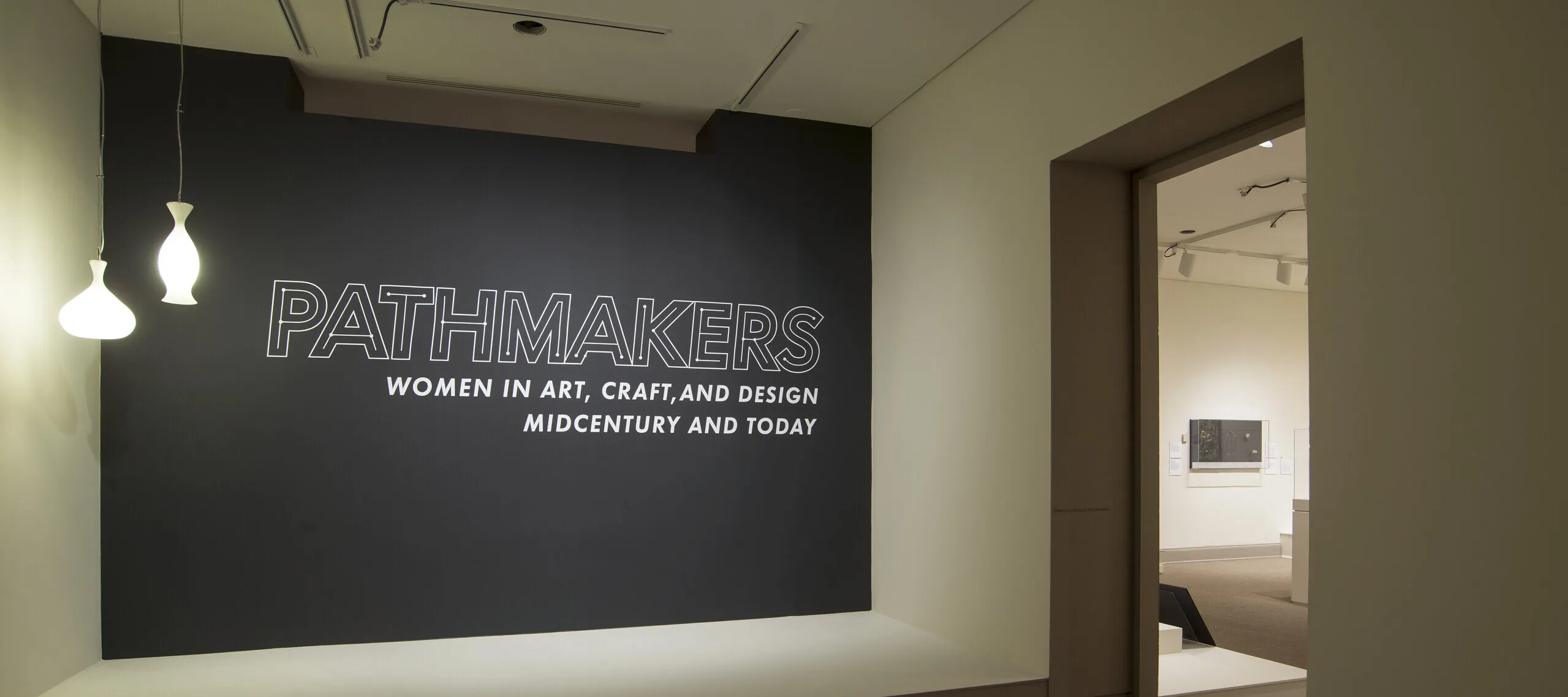 View of a gallery space. On a black wall, it says " Pathmakers: Women in Art, Craft, and Design, Midcentury and Today" in big, white letters, Two pendant lights are hanging from the ceiling to the left.