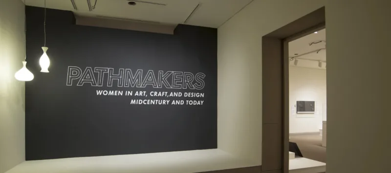 View of a gallery space. On a black wall, it says " Pathmakers: Women in Art, Craft, and Design, Midcentury and Today" in big, white letters, Two pendant lights are hanging from the ceiling to the left.