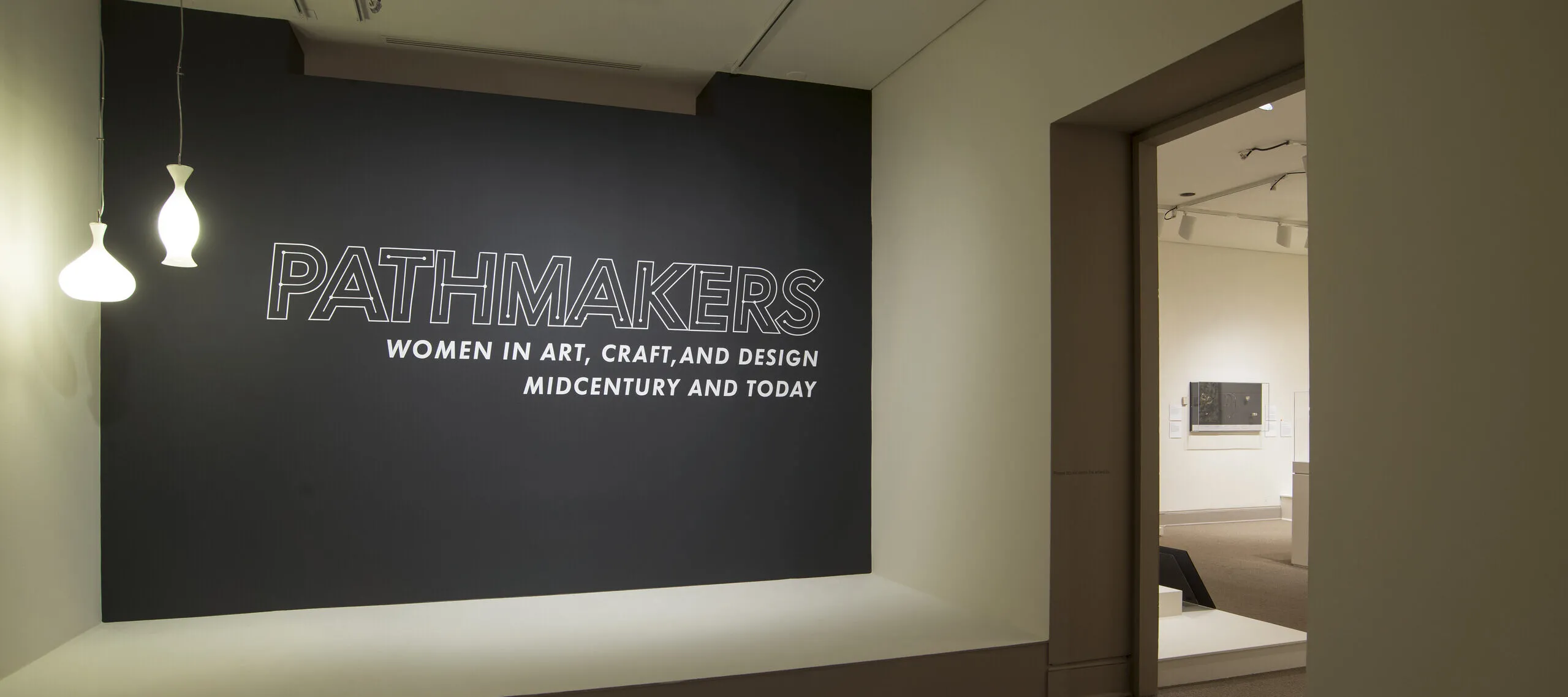 View of a gallery space. On a black wall, it says " Pathmakers: Women in Art, Craft, and Design, Midcentury and Today" in big, white letters, Two pendant lights are hanging from the ceiling to the left.