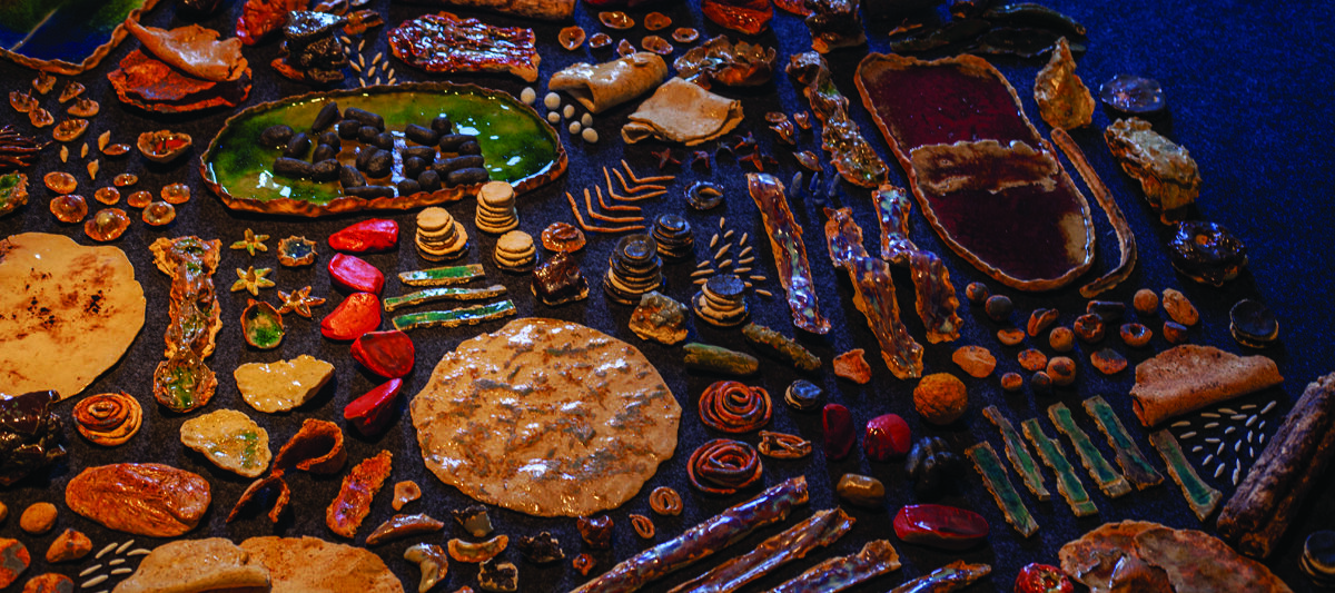 A sculptural arrangement of small, highly-detailed foods made out of ceramic are spaced evenly on the floor in the shape of a circle. The foods are glossy, with lots of textures and warm colors.