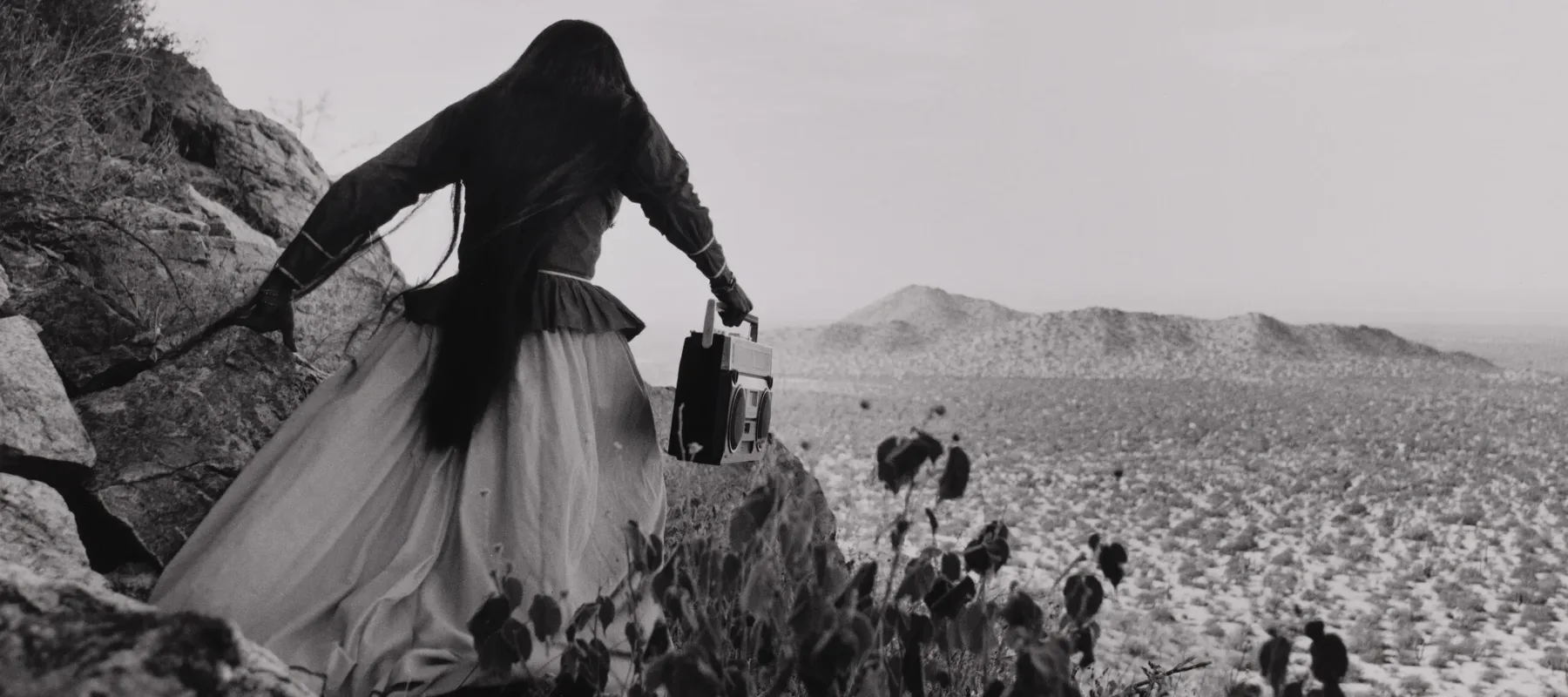 A black-and-white photograph shows the back of a woman as she crests a rocky path above a vast desert landscape beneath an expansive sky. Her traditional, ethnic full skirt, long-sleeved blouse, and long, straight, dark hair contrasts with the modern portable stereo she carries.