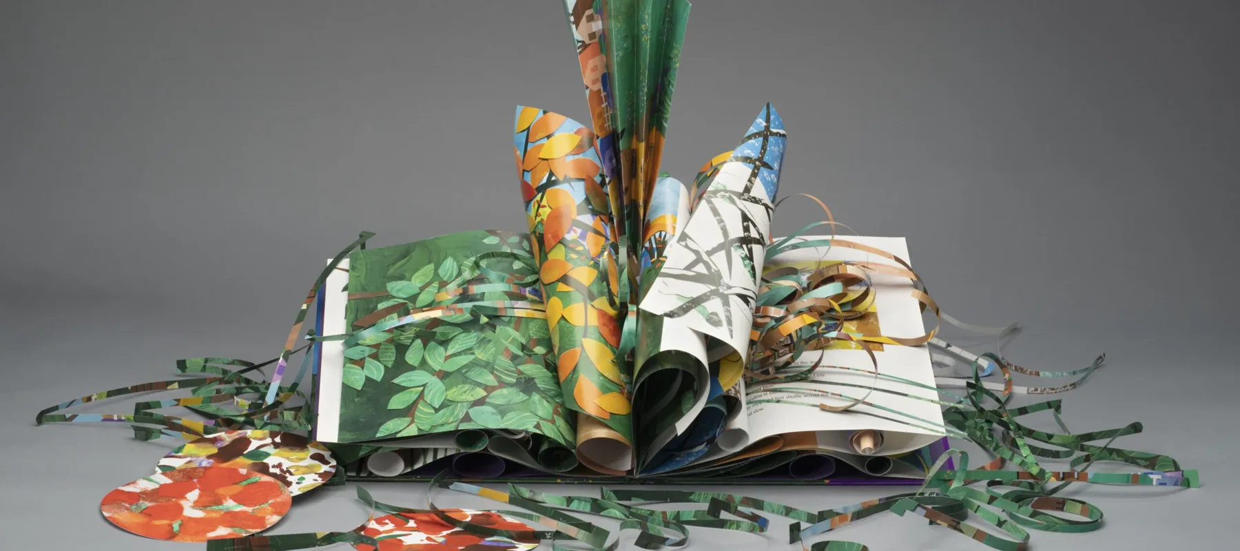 An artist's book made of colorful green, yellow, brown, orange, and red paper. Several of the pages are curled and folded. Other paper is cut into strips and circles and displayed around the book.