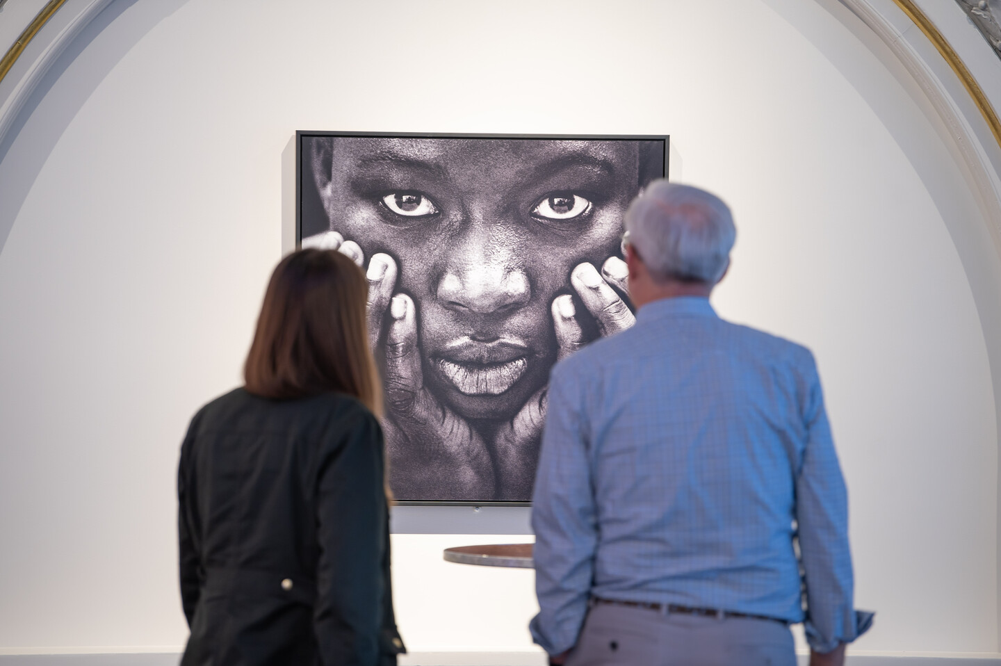 Two museum visitors are viewing an artwork. The artwork is a black-and-white close-up portrait of a woman's face. She has a dark skin tone and is holding her face in her hands.