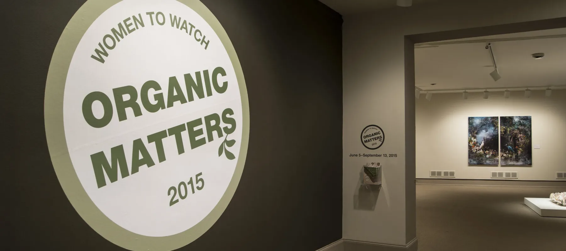 View of a gallery space. In big letters green , the text on the wall reads "Women to Watch: Organic Matters, 2015."