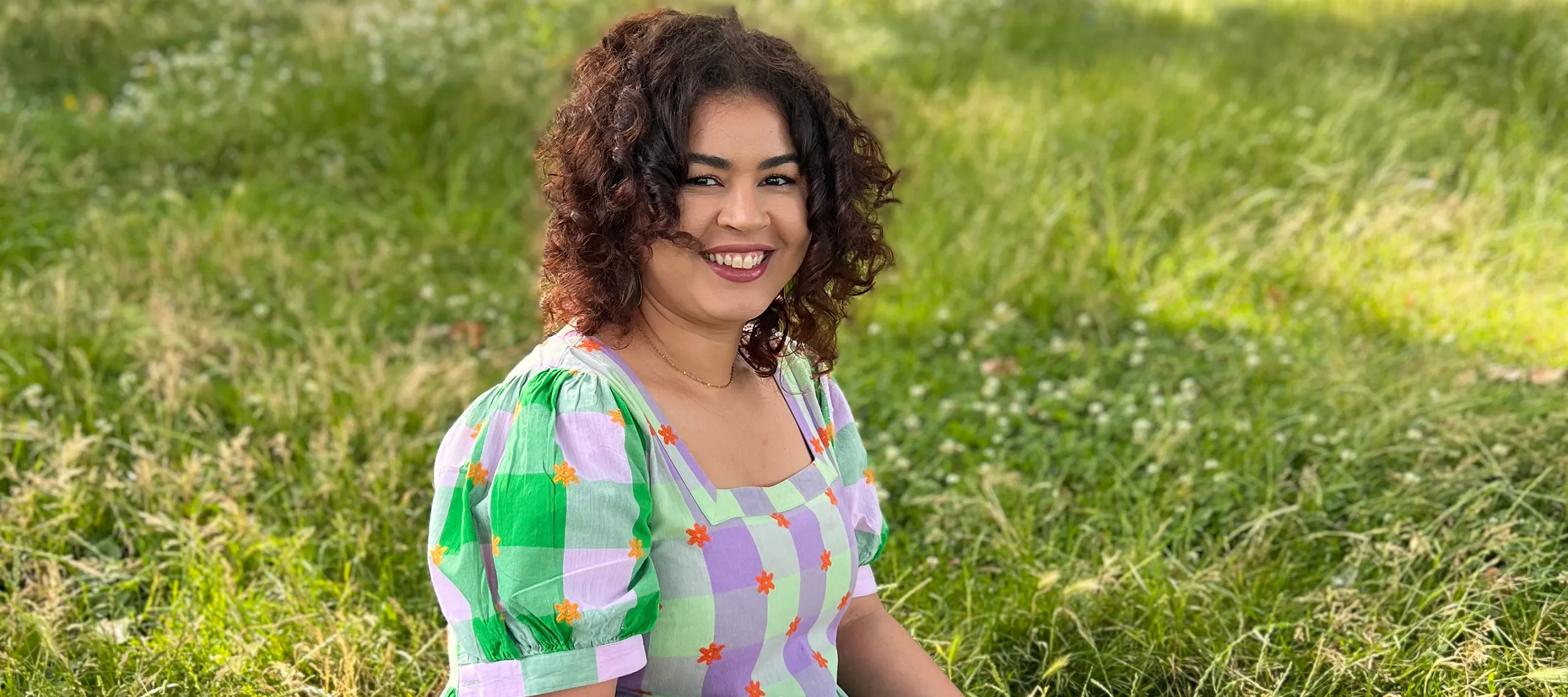 A young woman with olive skin tone and brown curly hair sits in green grass. She wears a floral and checkered long dress and holds a tablet and small pen. She smiles at the camera.