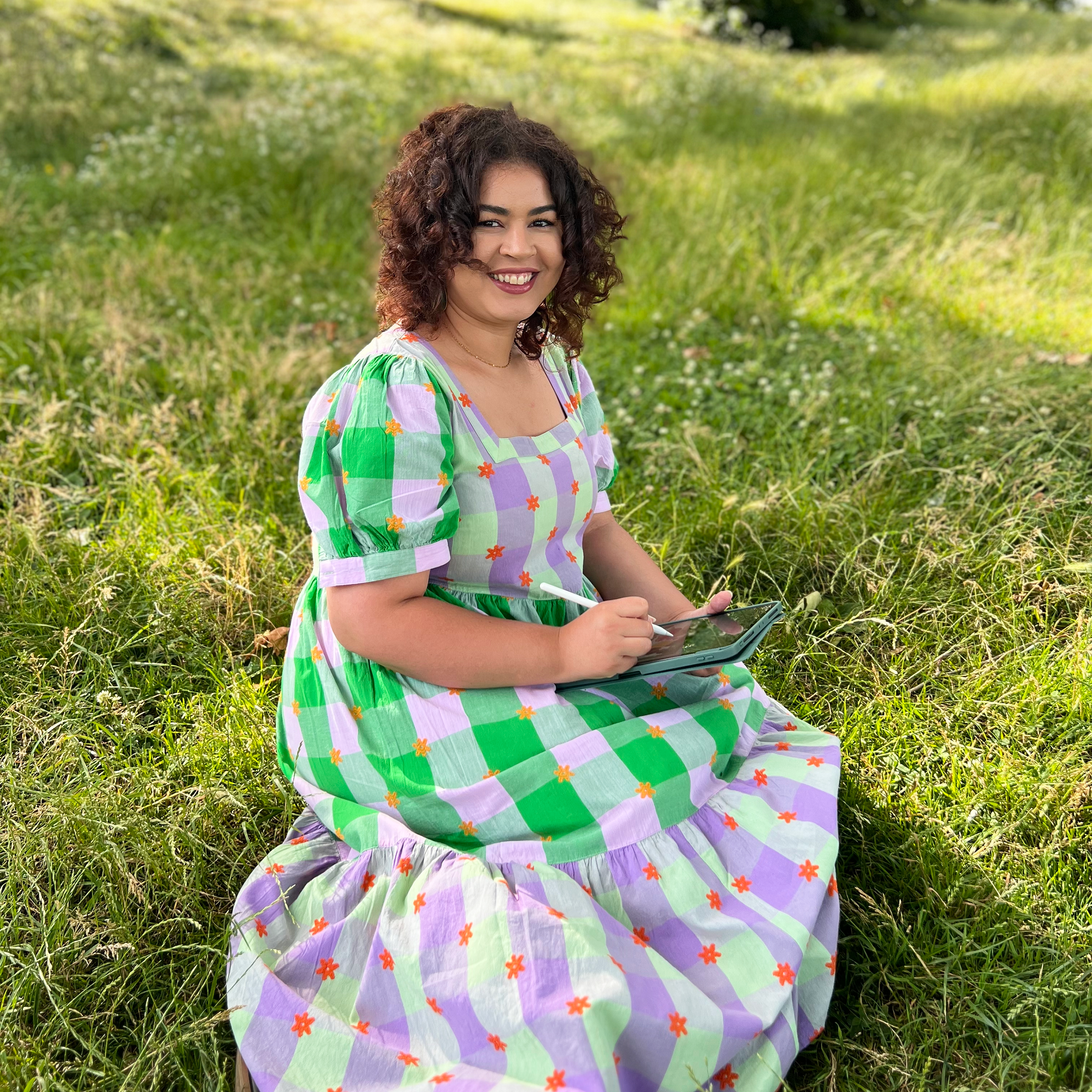 A young woman with olive skin tone and brown curly hair sits in green grass. She wears a floral and checkered long dress and holds a tablet and small pen. She smiles at the camera.