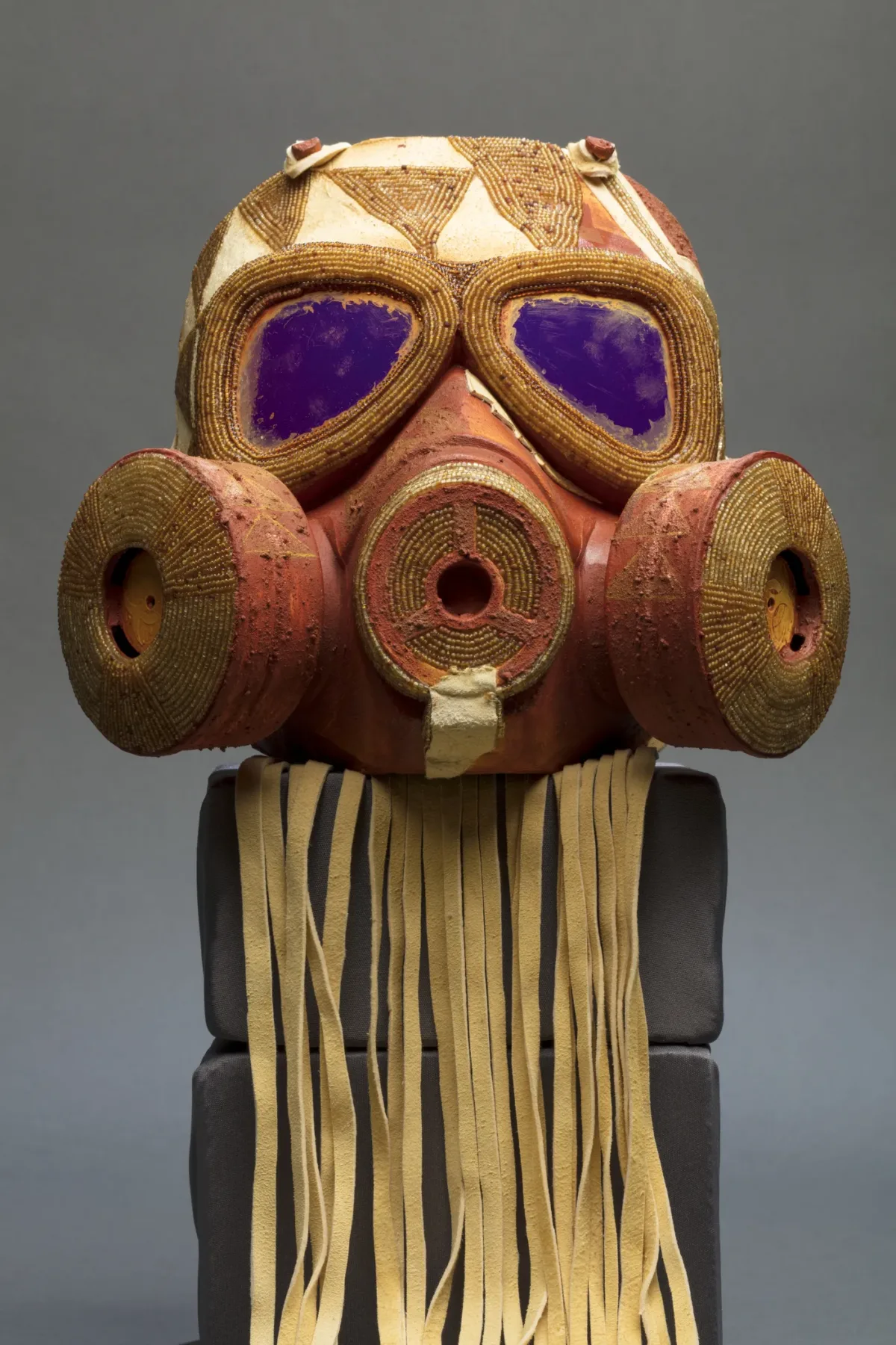 An orange and yellow gas mask embellished with gold beading sitting atop two stacked bricks with beige fringe hanging from its base.