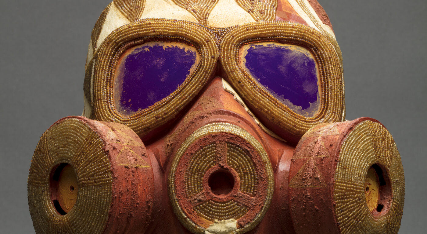 An orange and yellow gas mask embellished with gold beading sitting atop two stacked bricks with beige fringe hanging from its base.