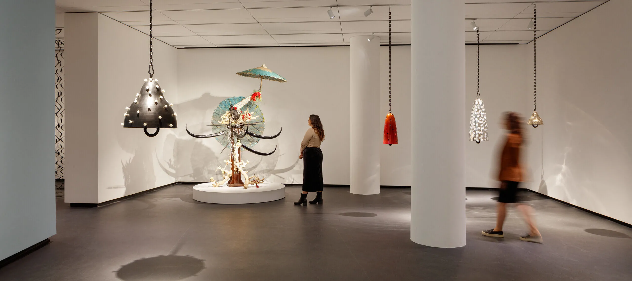 A modern museum gallery is photographed at a wide angle. Visitors observe large sculptures, several of which hang from the ceiling.