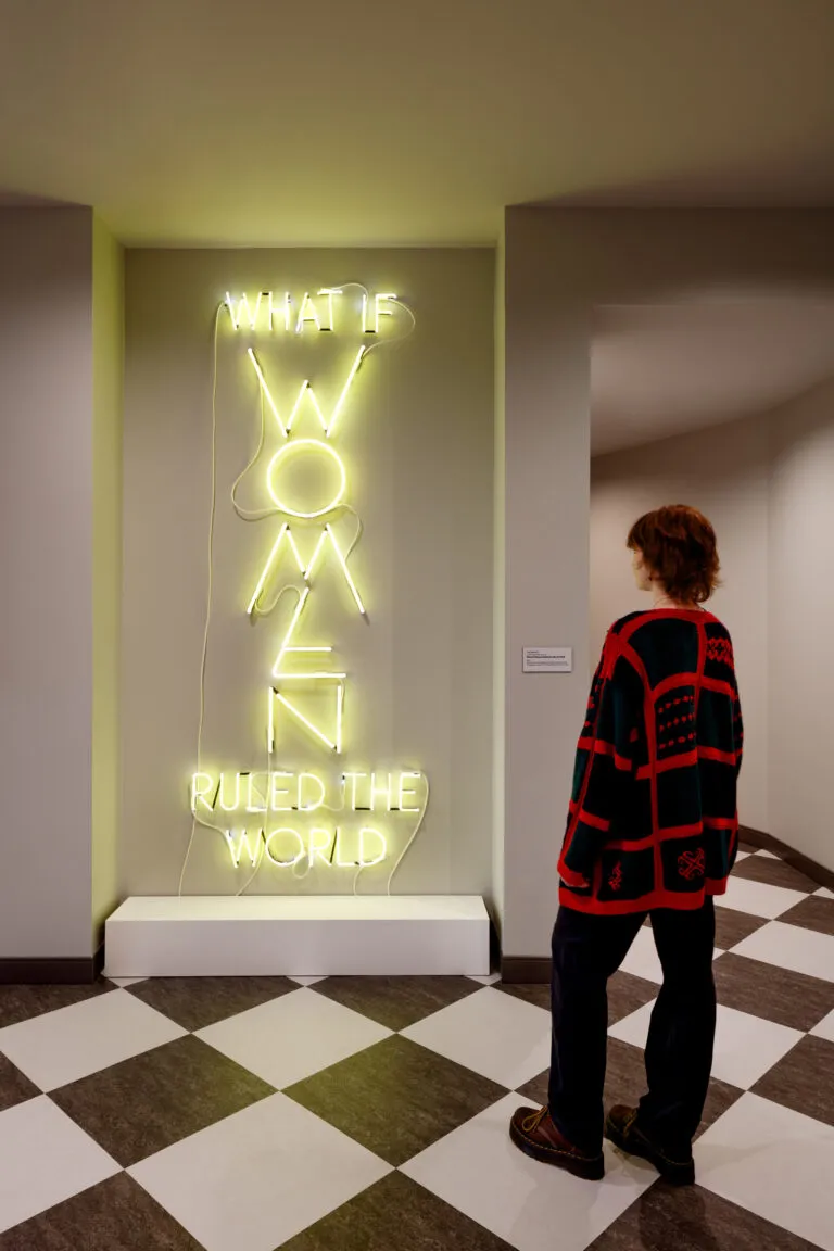 A museum visitor looks at a neon artwork hanging on a wall. The phrase "What if Women Ruled the World" appears sculpted with neon yellow tube lights, in all capital letters. The letters of “WOMEN” are larger than the rest and arranged vertically.