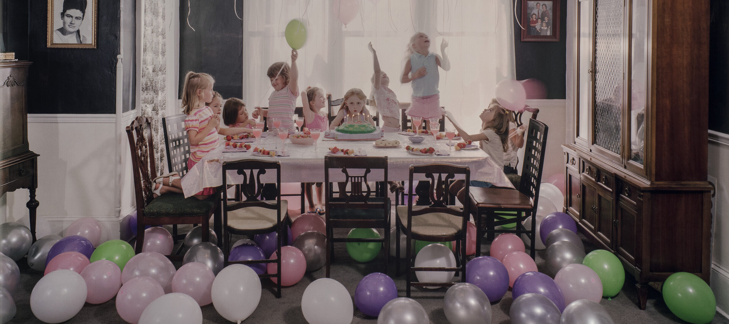 Green, purple, silver, white and pink balloons litter the floor and cover the ceiling of a navy-walled family dining room. In the middle of the room, a table is set with a pink table cloth. In the center of the table, a young girl blows out the candles on her birthday cake while looking straight ahead. Around her, nine other young girls jump, spin, and play.