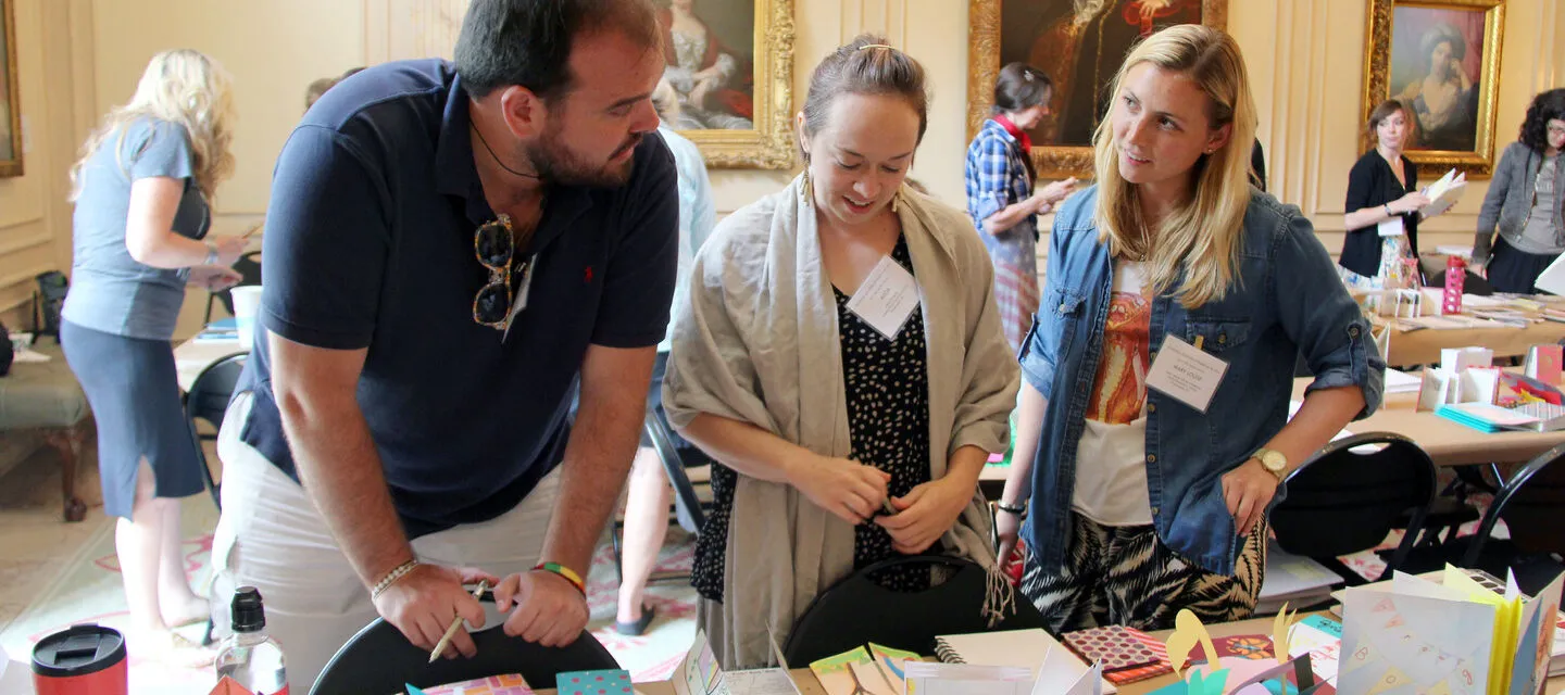 Three people with light skin tones stand in front of a table full of artists' books look at each other and chat.