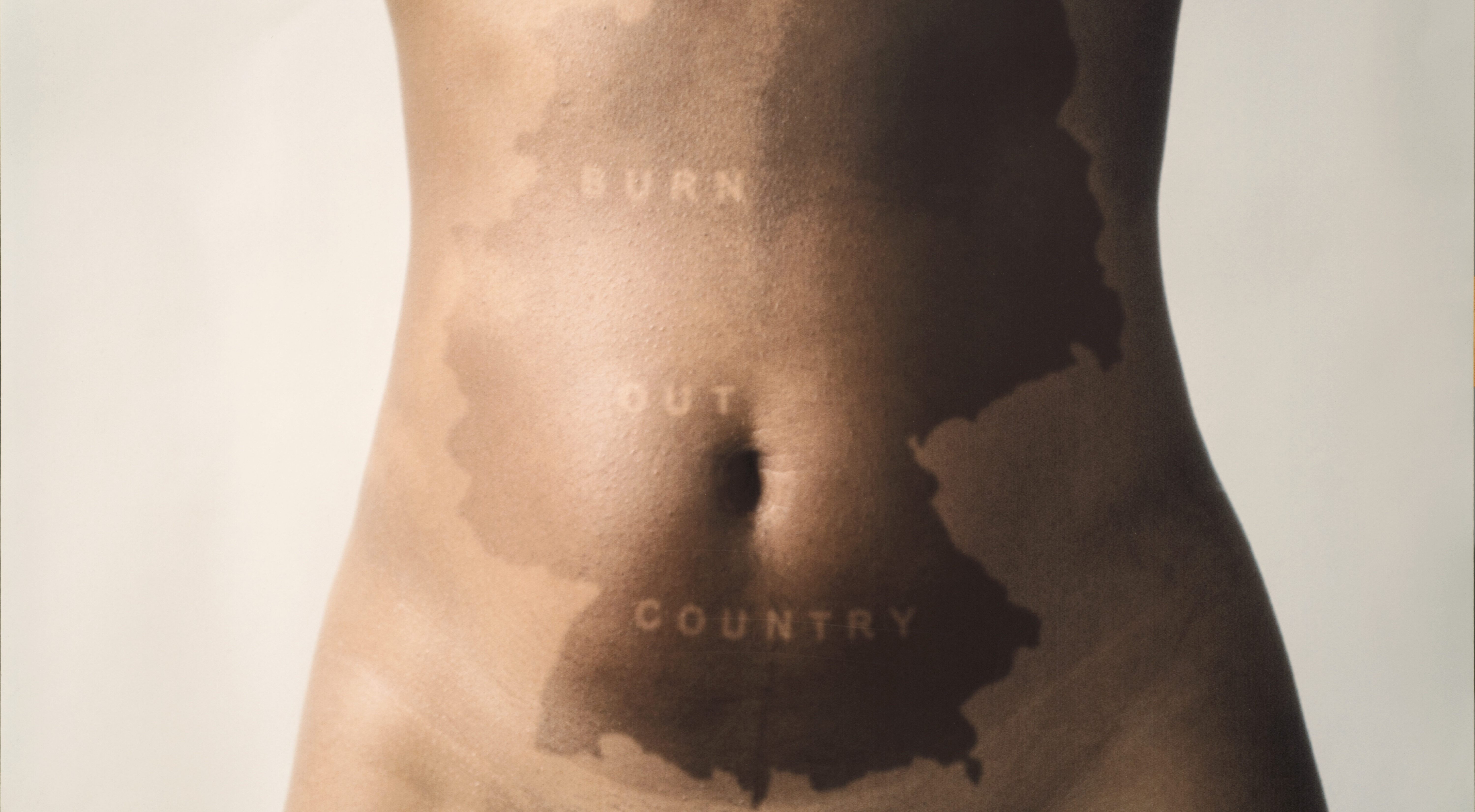 The nude torso of a woman is pictured from sternum to hip. In the center of her stomach a darker patch of skin makes the outline of Germany. Through the darker skin, light letter run diagonally that read 