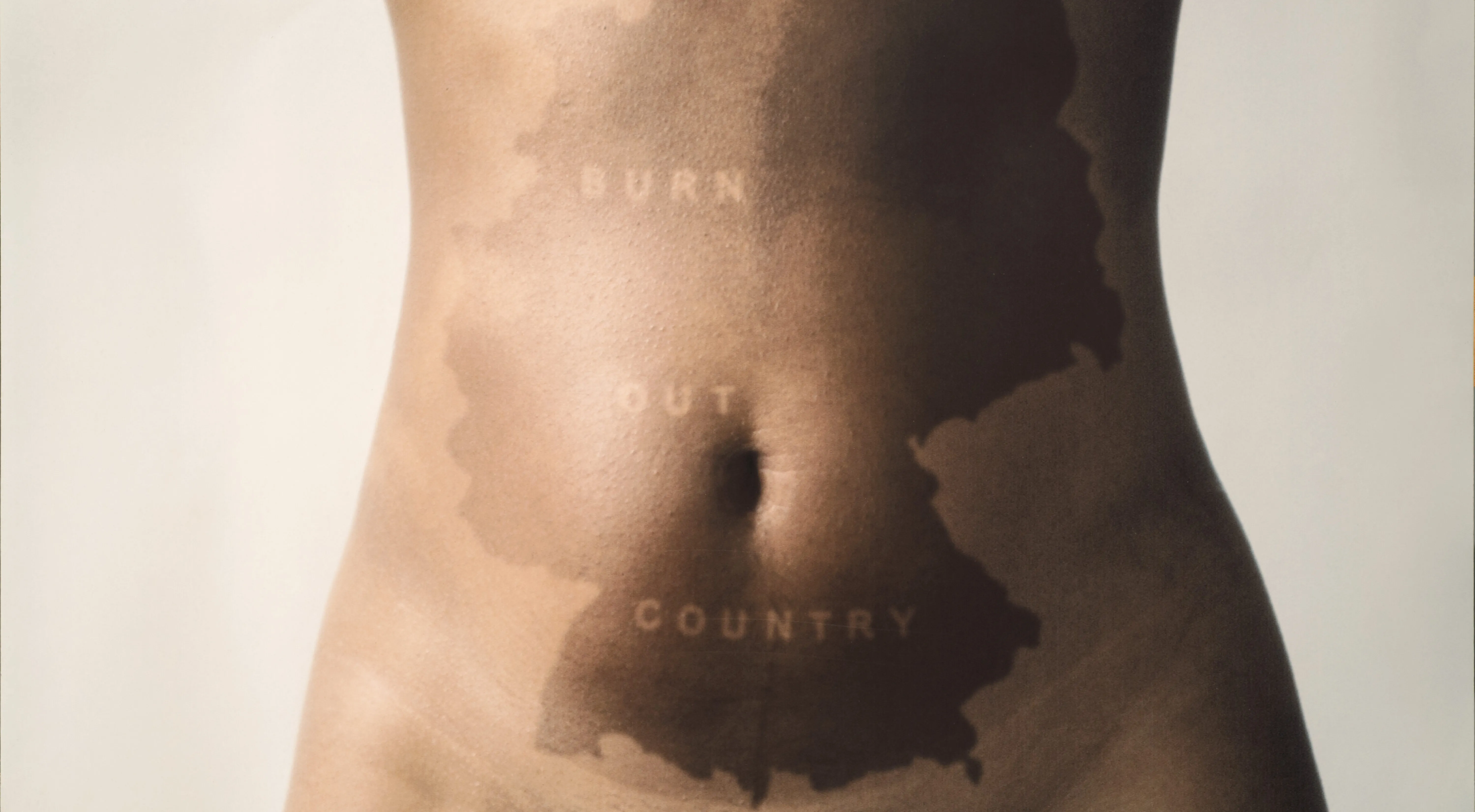 The nude torso of a woman is pictured from sternum to hip. In the center of her stomach a darker patch of skin makes the outline of Germany. Through the darker skin, light letter run diagonally that read "Burn Out Country".