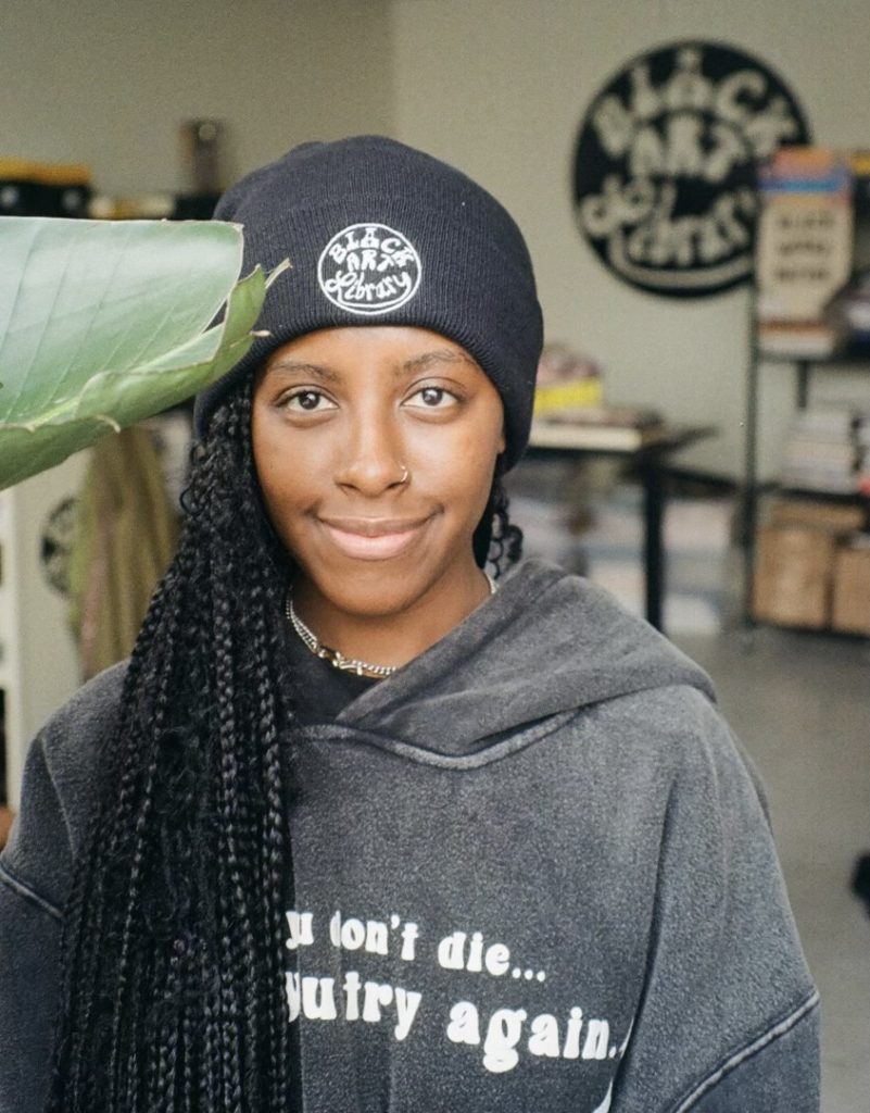 A portrait of a woman with a dark skin tone wearing a beanie and a gray hoodie standing next to a large plant. She has long, braided black hair and is smiling towards the camera. 