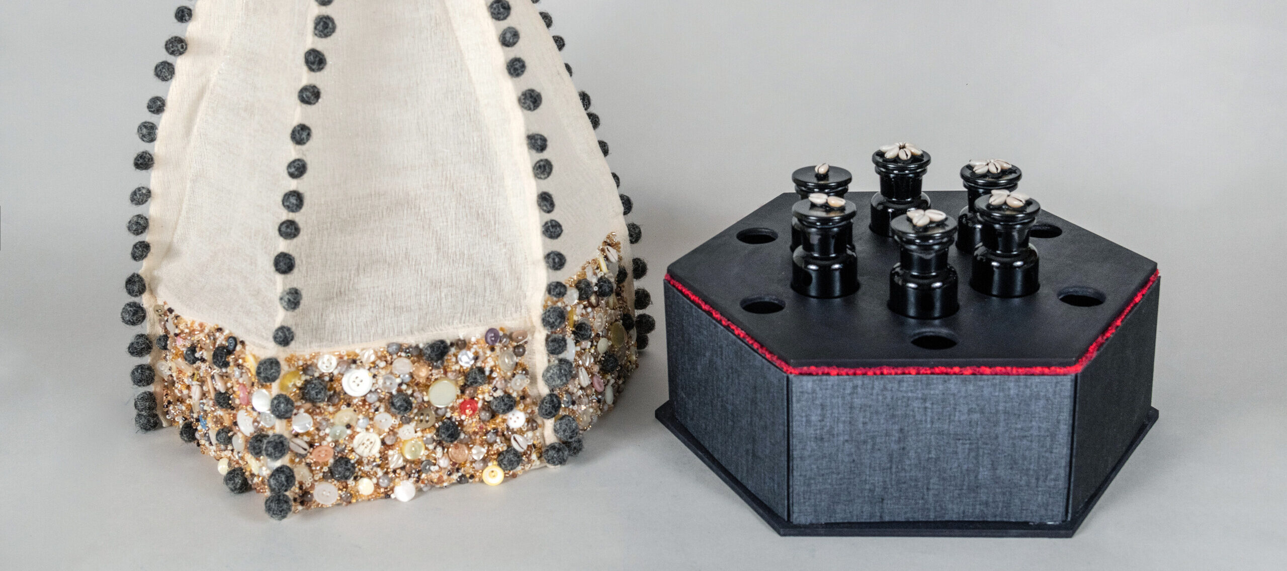Two parts of an artist's book. One part is a black hexagonal box covered in cowry shells, small black bottles, and red beads. The other part is made of white fabric and covered in balls of human hair, buttons, and beads.