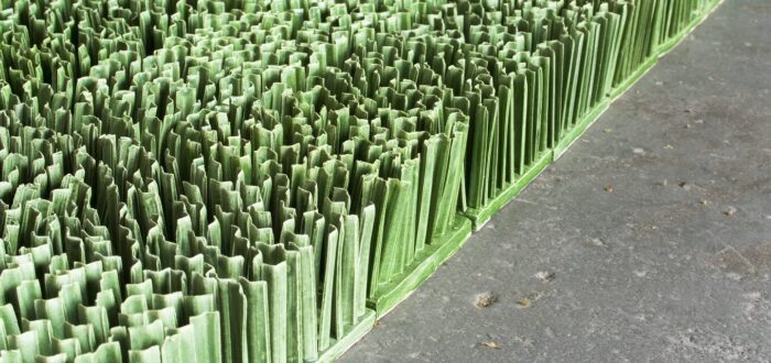 Detail photograph of ceramic sculpture made to look like a patch of lawn. Individual squares consisting of multiple upright blades of porcelain grass, glazed green, fit together to form a lush rectangular field of grass.