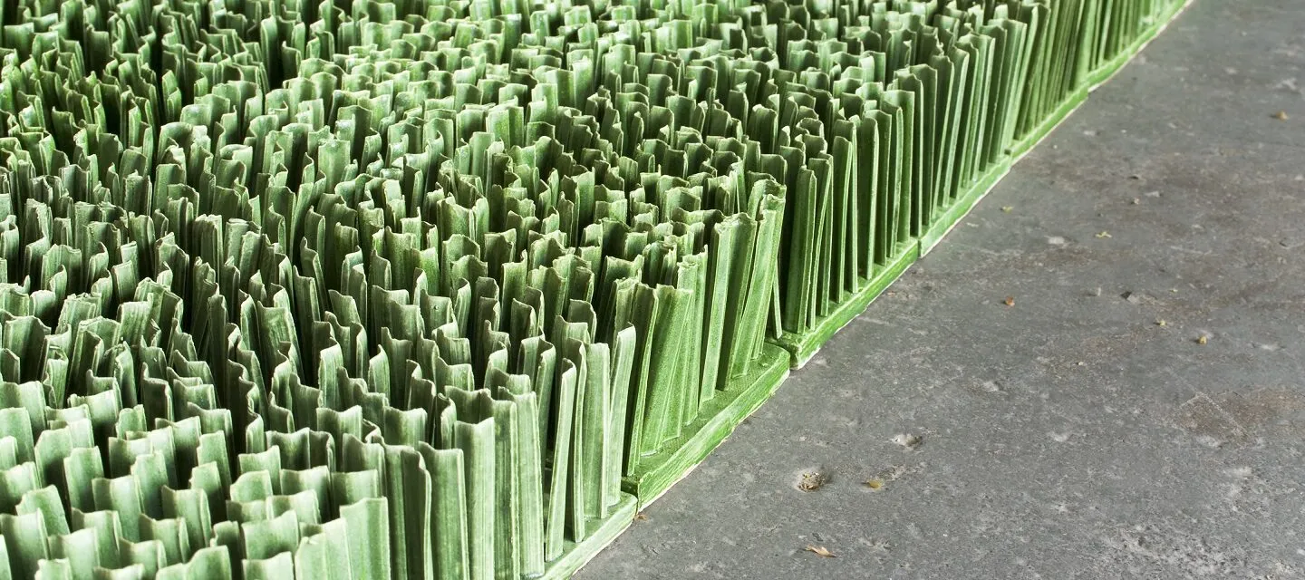 Detail photograph of ceramic sculpture made to look like a patch of lawn. Individual squares consisting of multiple upright blades of porcelain grass, glazed green, fit together to form a lush rectangular field of grass.