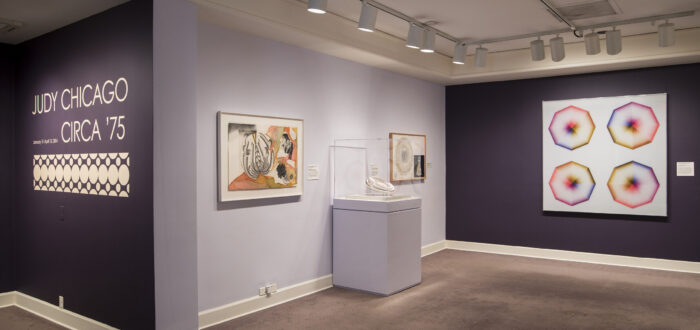 Installation view of a gallery space with purple walls. Several colorful art pieces are hanging on the wall. On one wall, the text says: Judy Chicago, circa '75.