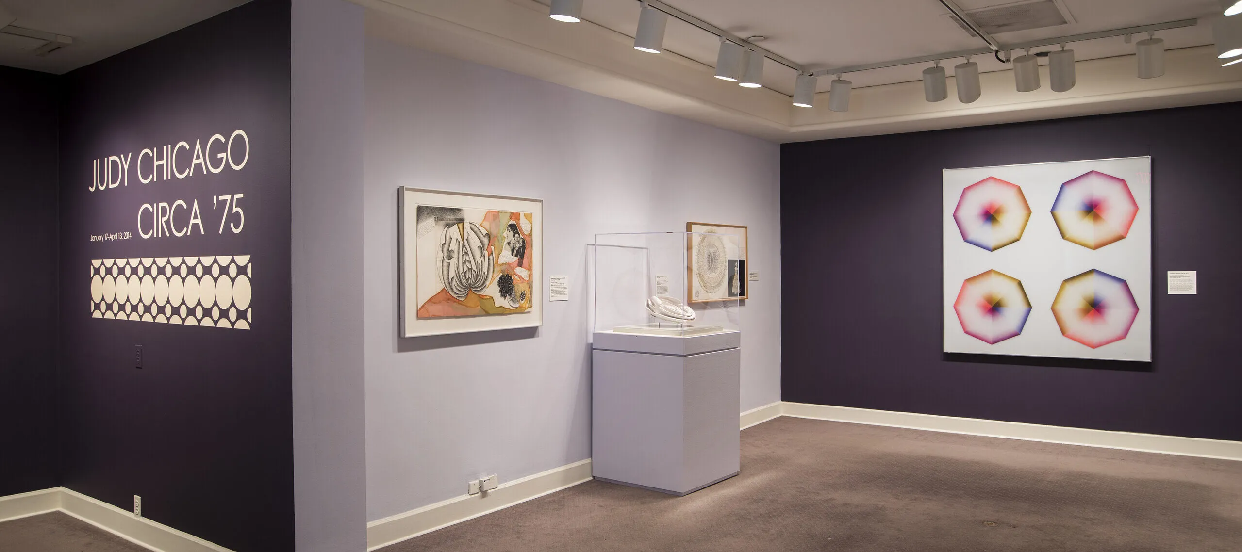 Installation view of a gallery space with purple walls. Several colorful art pieces are hanging on the wall. On one wall, the text says: Judy Chicago, circa '75.