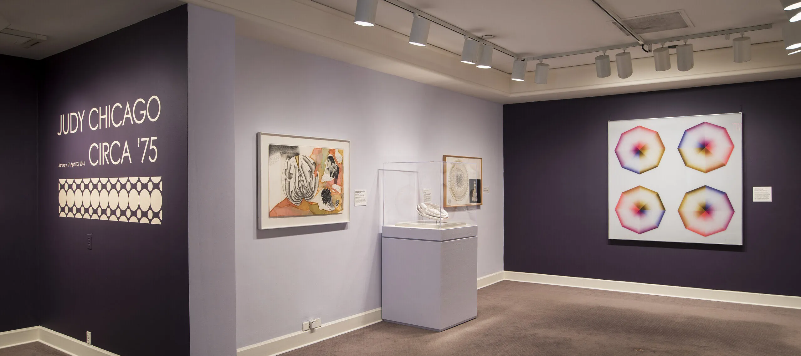 Installation view of a gallery space with purple walls. Several colorful art pieces are hanging on the wall. On one wall, the text says: Judy Chicago, circa '75.