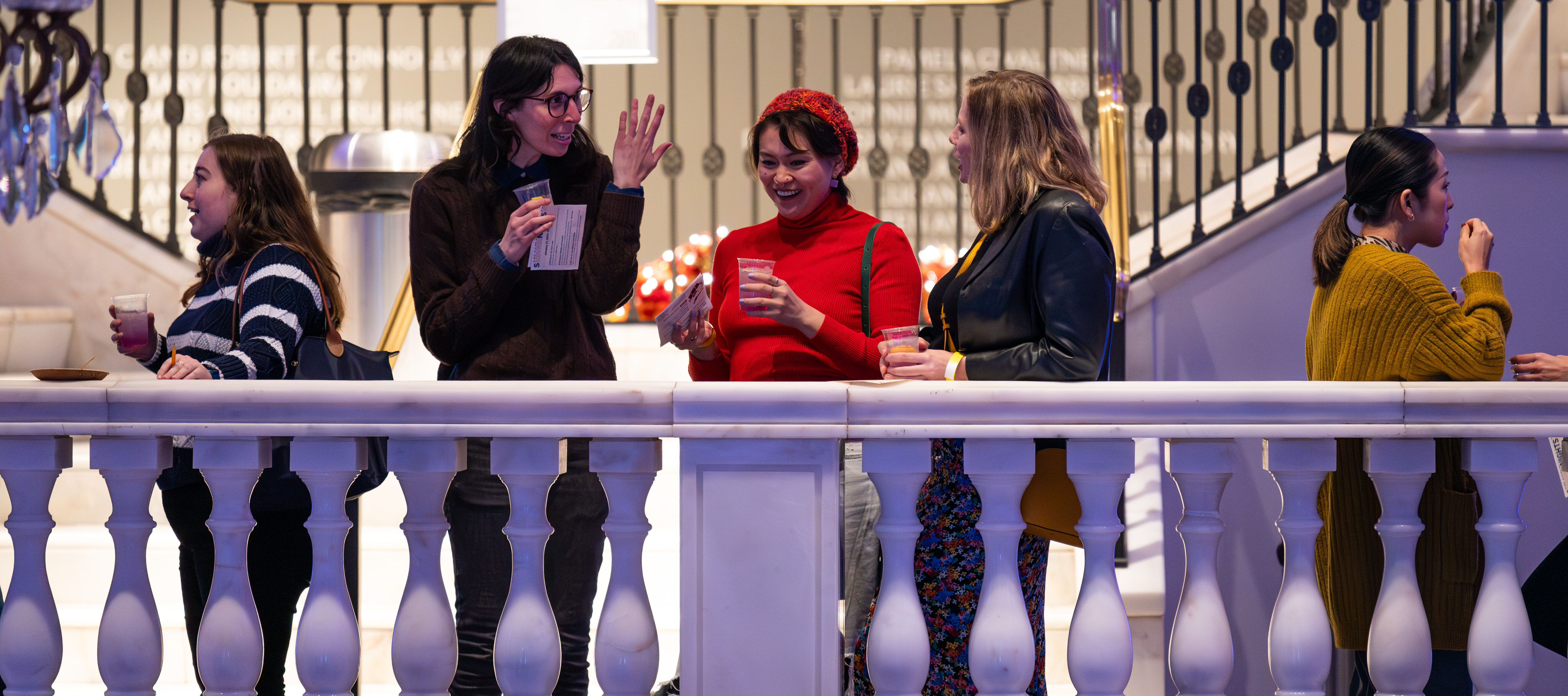 Five women talking and drinking beverages on a balcony in a museum.
