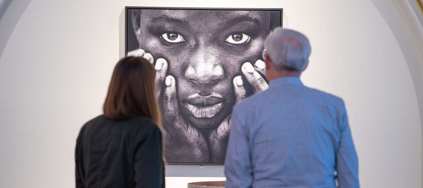 Two museum visitors are viewing an artwork. The artwork is a black-and-white close-up portrait of a woman's face. She has a dark skin tone and is holding her face in her hands.