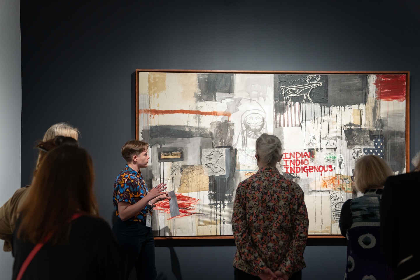 An person stands in front of a painting in a museum surrounded by a group of people. They are holding a paper with text on it. They have a light skin tone, short hair, and wear a colorful shirt with abstract patterns.