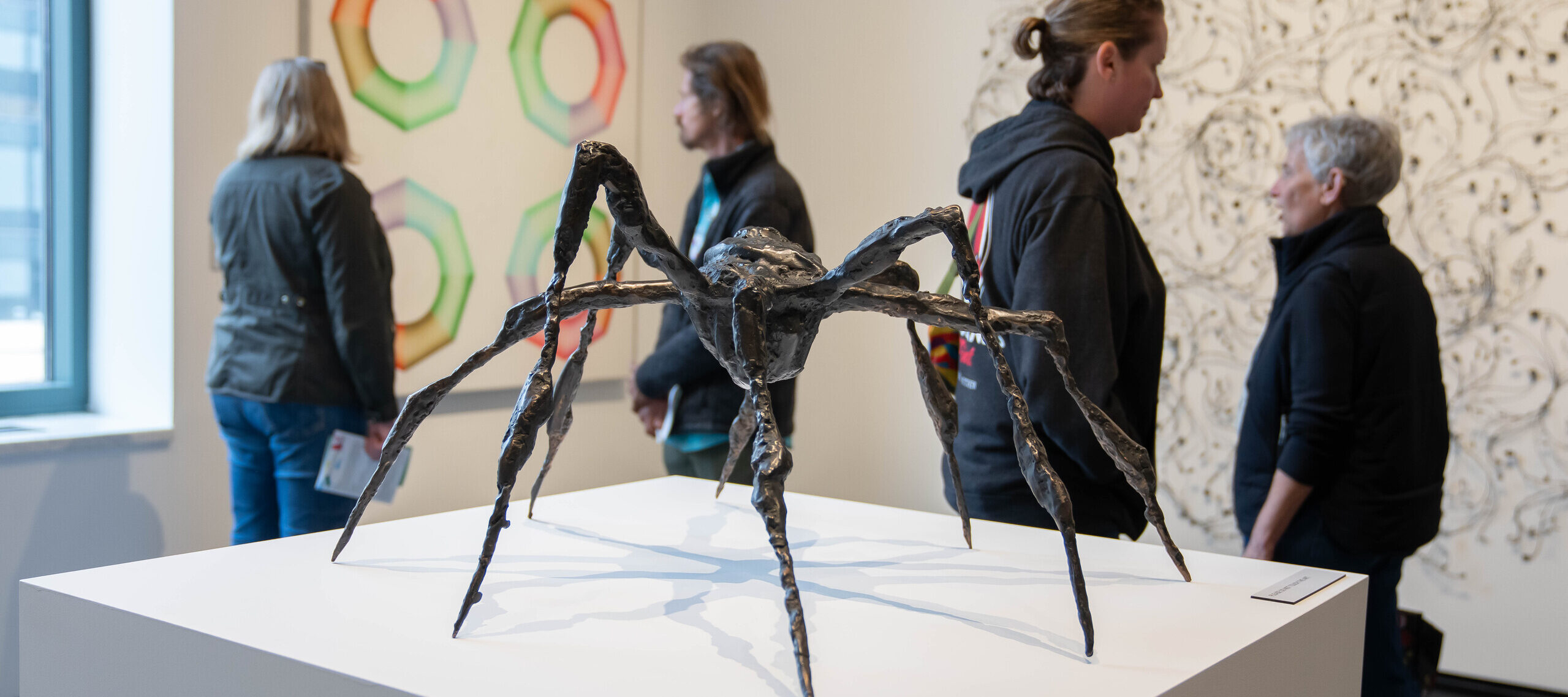 A bronze sculpture of a spider with arched, spindly legs sits on a white pedestal in a museum gallery. The spider appears to be frozen mid-step with some legs raised higher than the others. In the background, museum visitors observe artworks on the walls.