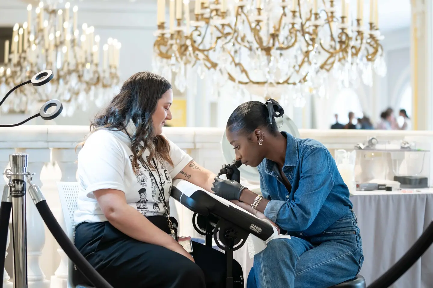 A woman with a medium skin tone and wavy, brunette hair is getting a tattoo. The tattoo artist has a dark skin tone and is wearing an all-denim outfit. They are seated in front of a baroque balustrade and a golden chandelier.