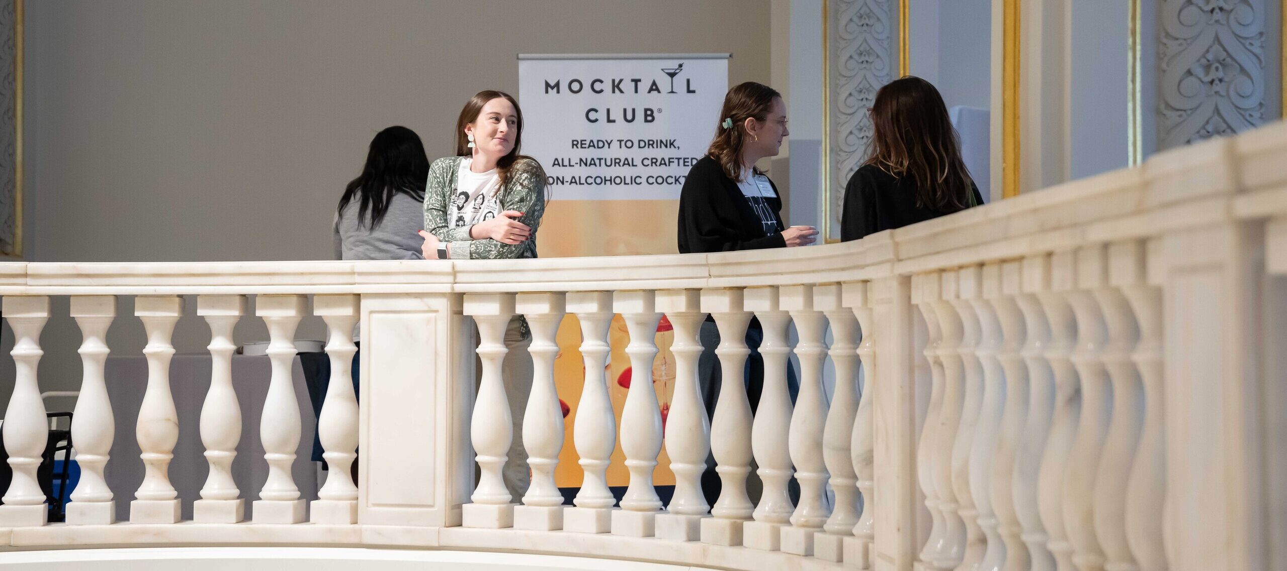 Four women are standing by a balustrade. One of the women is leaning against it. She has a light skin tone, long hair, and is wearing a white graphic tee. She is standing in front of a sign that says 