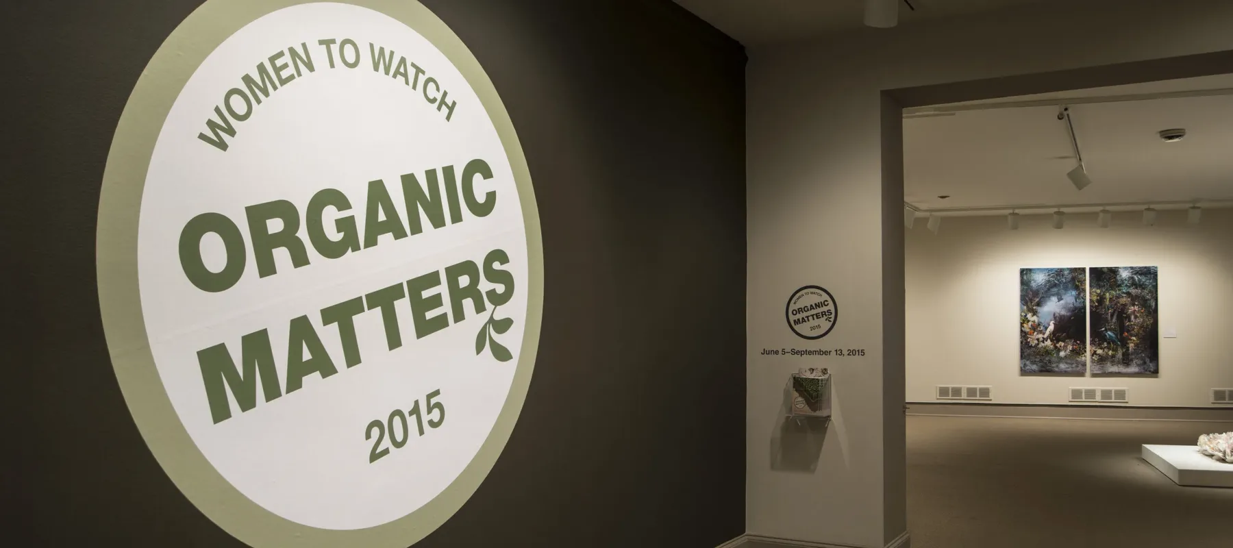 View of a gallery space. In big letters green , the text on the wall reads "Women to Watch: Organic Matters, 2015."