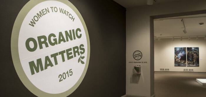 View of a gallery space. In big letters green , the text on the wall reads "Women to Watch: Organic Matters, 2015."