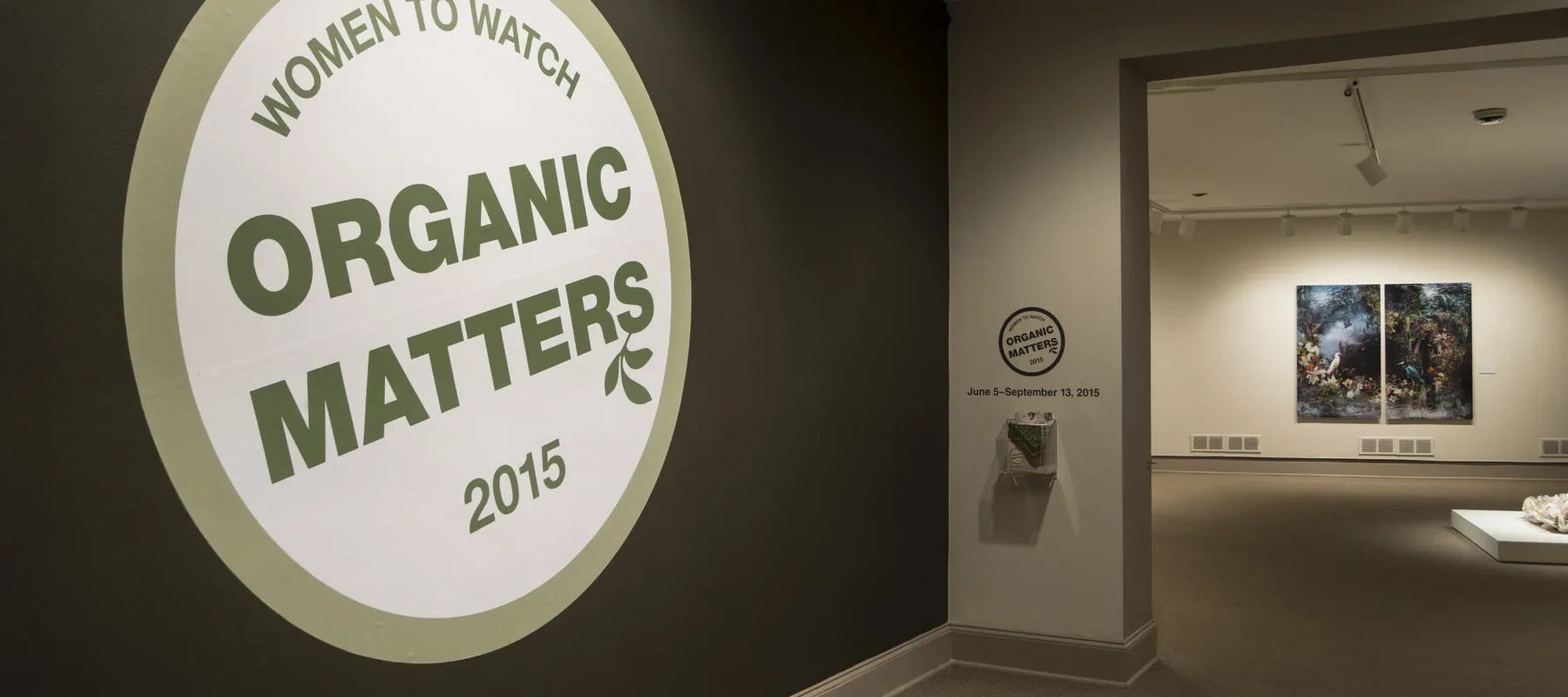 View of a gallery space. In big letters green , the text on the wall reads "Women to Watch: Organic Matters, 2015."