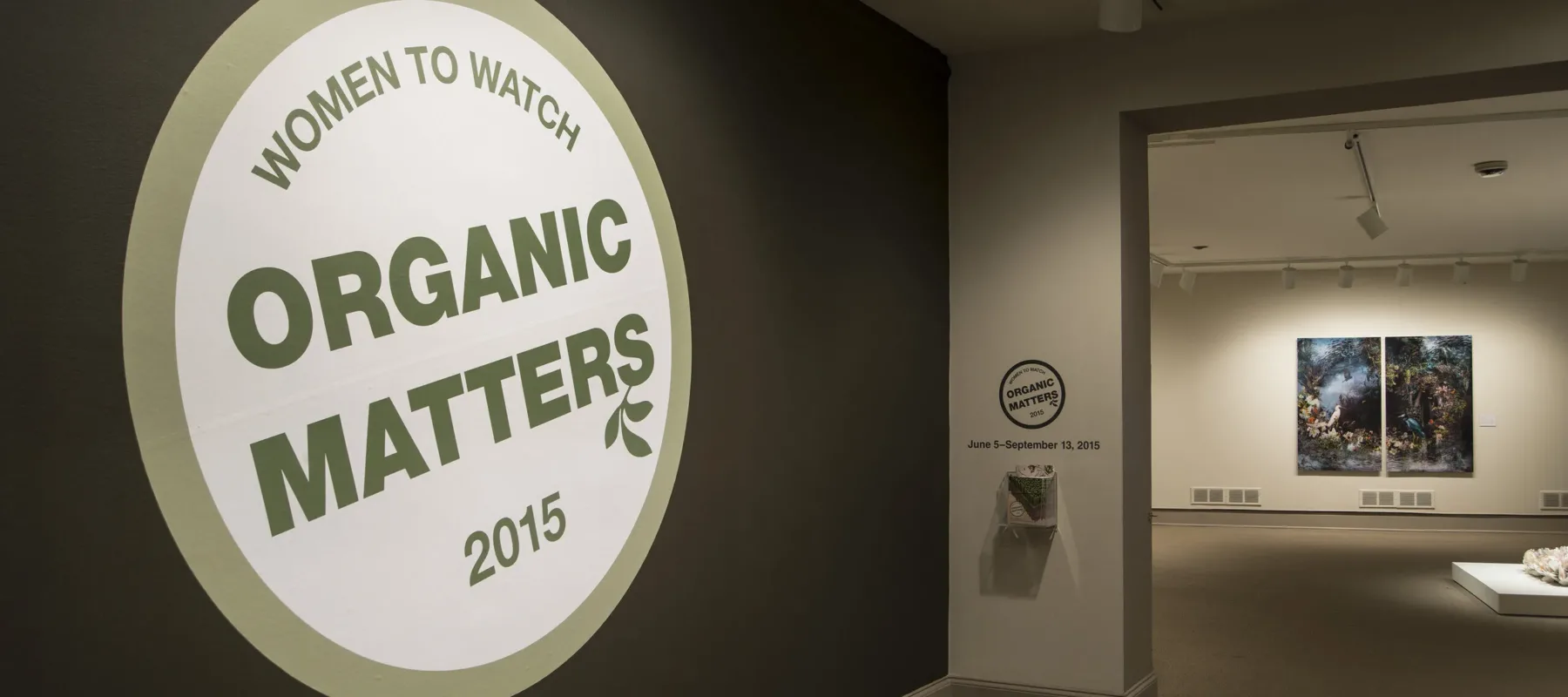 View of a gallery space. In big letters green , the text on the wall reads "Women to Watch: Organic Matters, 2015."