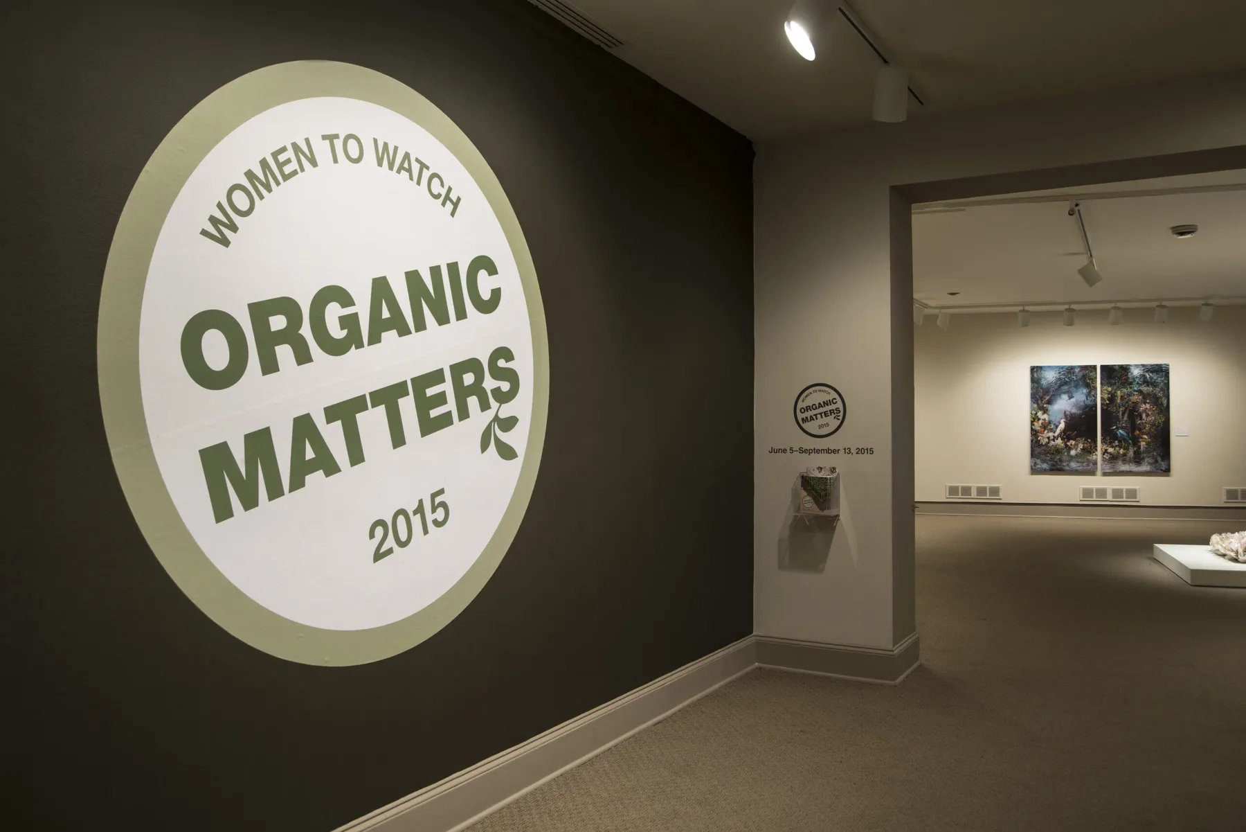 View of a gallery space. In big letters green , the text on the wall reads "Women to Watch: Organic Matters, 2015."