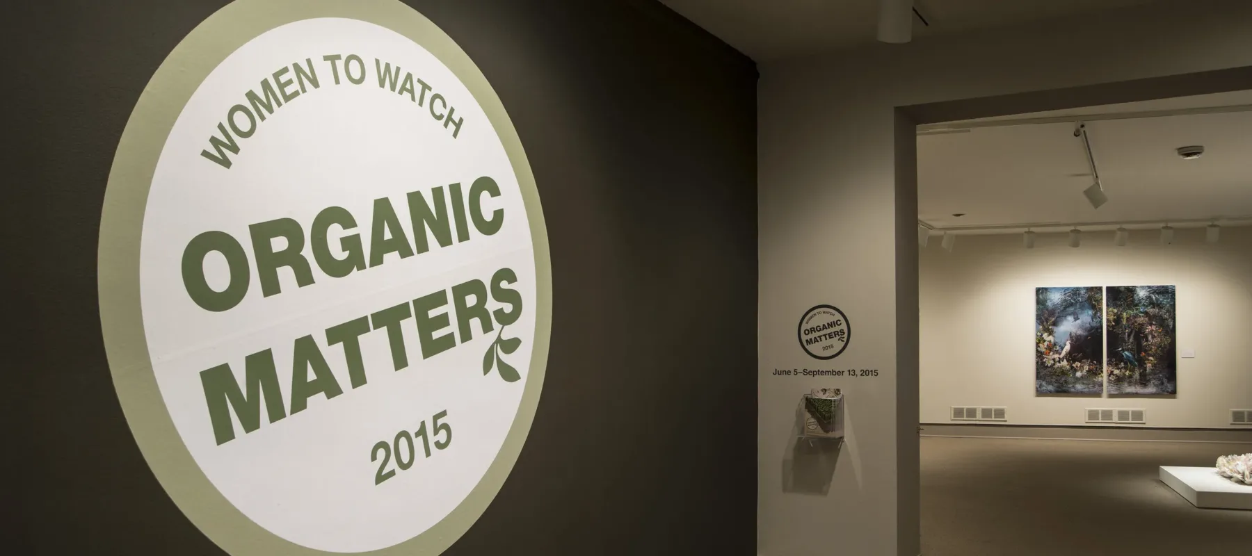 View of a gallery space. In big letters green , the text on the wall reads "Women to Watch: Organic Matters, 2015."